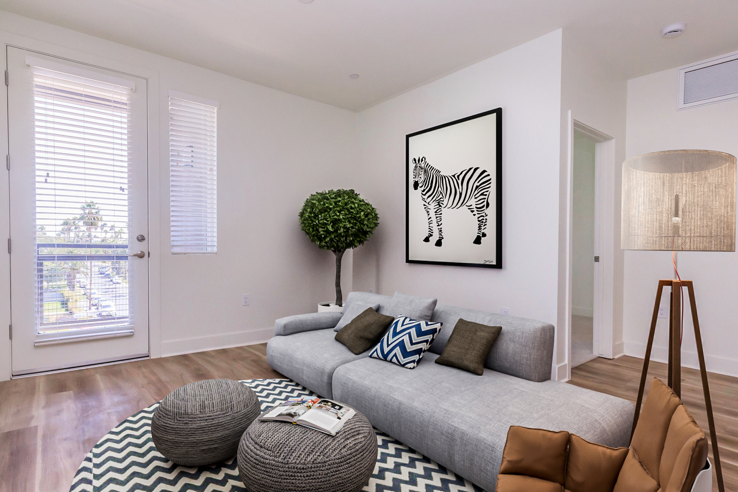 A modern living room featuring a gray sectional sofa with decorative pillows, a round patterned rug, two poufs, and a magazine on the floor. A large framed zebra artwork hangs on the wall, accompanied by a tall indoor plant and a stylish lamp. Natural light enters through large windows.