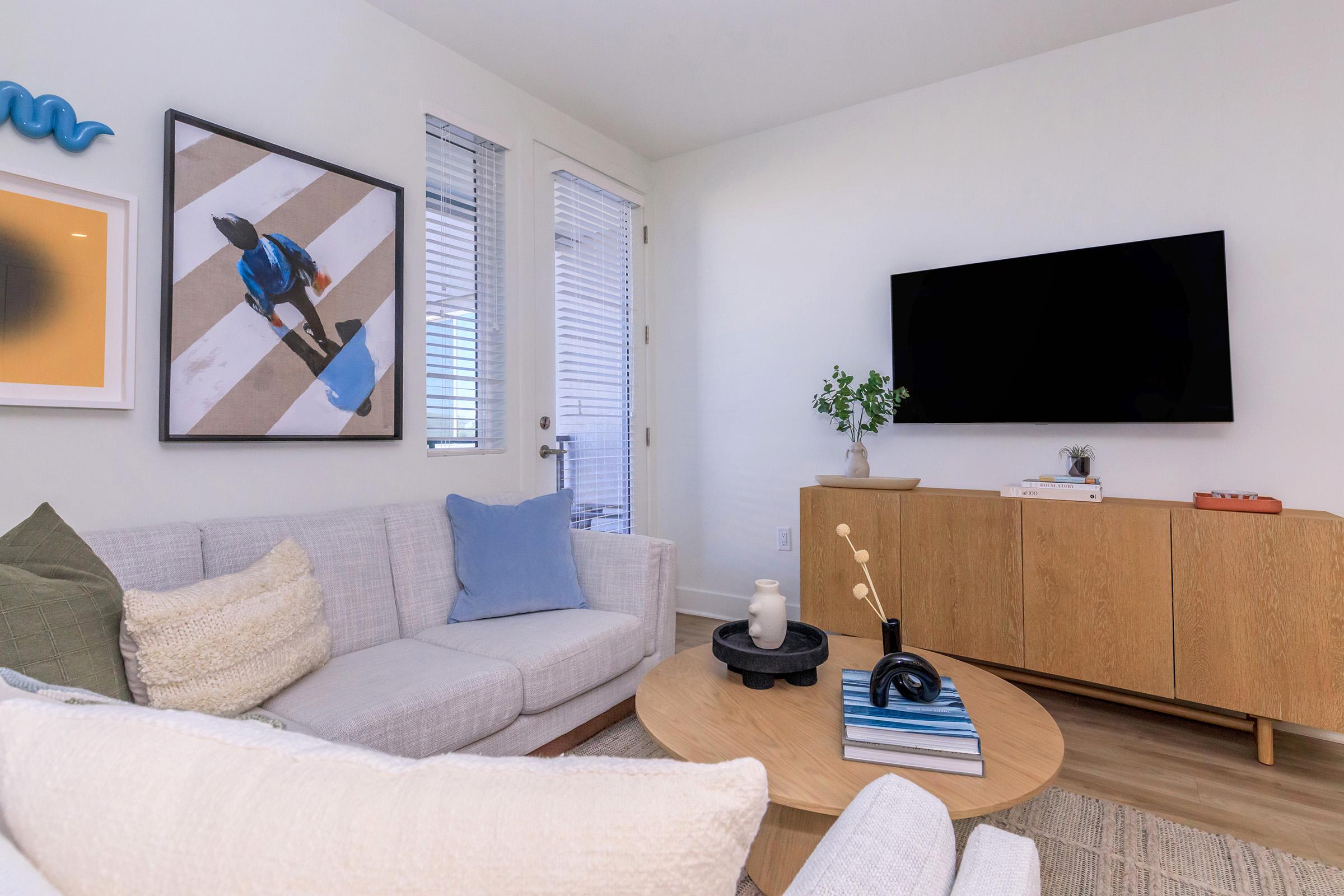 A modern living room featuring a light-colored sectional sofa with textured pillows, a round coffee table with decor, and a wooden TV stand. On the wall, there's a framed artwork of a person walking on a street. Large windows provide natural light, and a plant adds a touch of greenery.