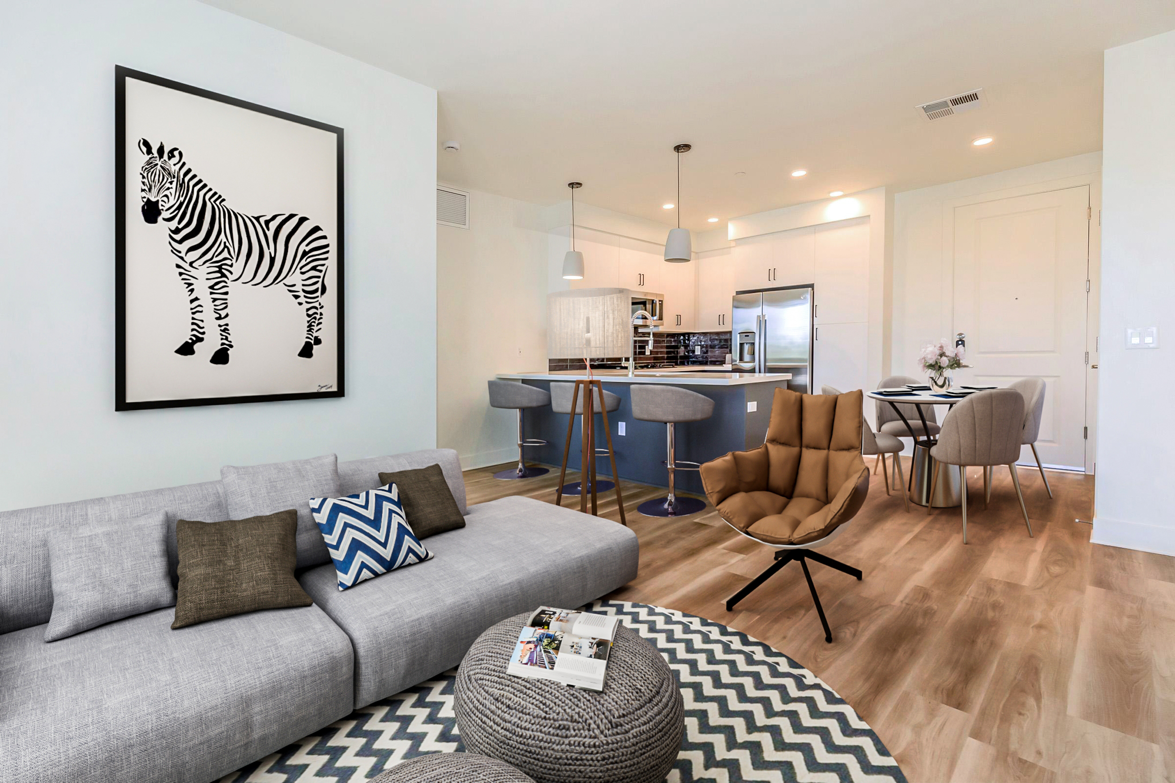 A modern living room featuring a gray sectional sofa with decorative pillows, a round area rug, and a coffee table. There is a large framed artwork of a zebra on the wall, along with a kitchen visible in the background. The kitchen has white cabinets, gray countertops, and barstool seating.