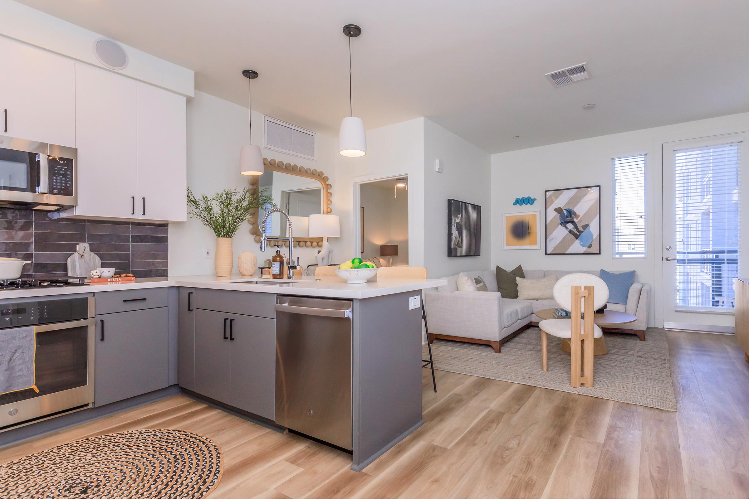 Modern kitchen with stainless steel appliances, gray cabinets, and a stylish backsplash. A living area features a light-colored sofa, a table, and decorative artwork. Large windows provide natural light, and there are plants and decorative elements enhancing the cozy atmosphere.