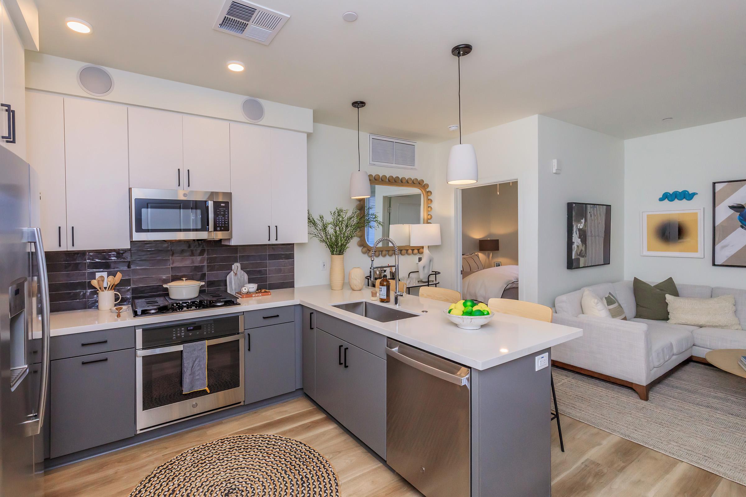 Modern kitchen featuring sleek, white cabinets, stainless steel appliances, and a gray island with a sink. In the background, a cozy living area with a light sofa, artwork on the walls, and decorative plants. Bright and airy atmosphere with natural light.