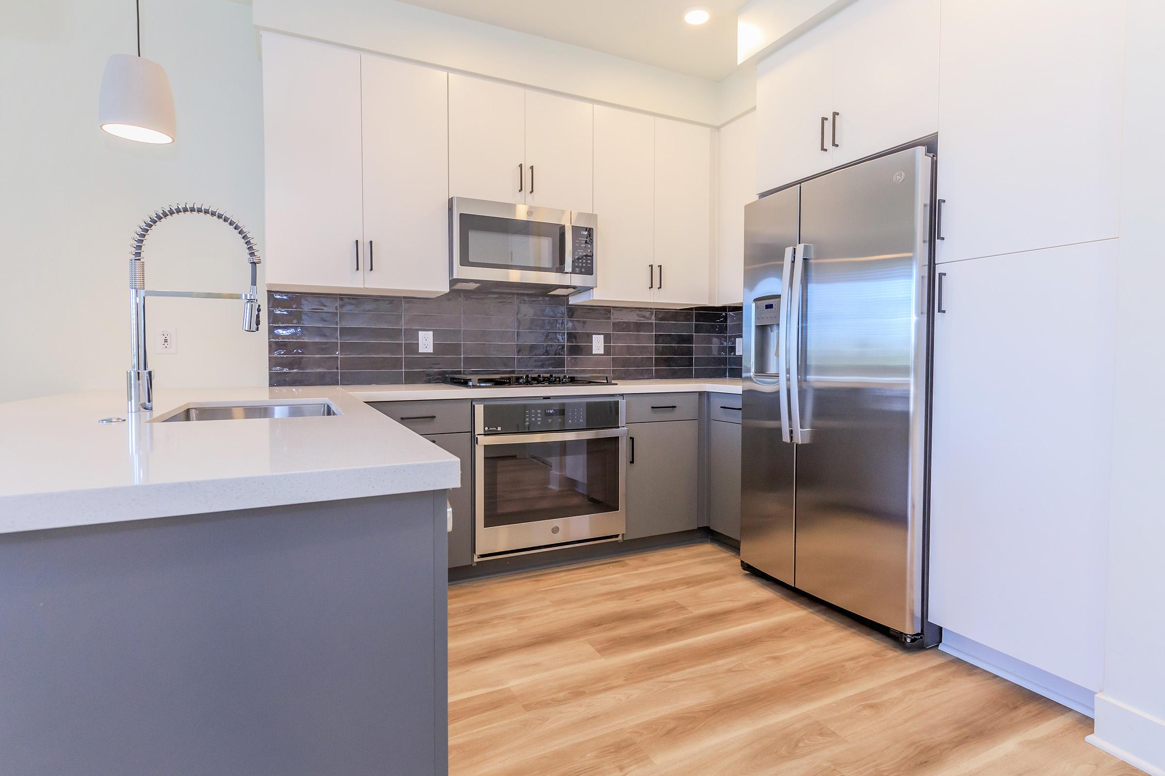 Modern kitchen featuring stainless steel appliances, including a microwave and refrigerator. The space has light gray cabinetry and a white countertop, with a sink and a stylish faucet. The floor is made of hardwood, and the walls are painted a light color, creating a bright and inviting atmosphere.