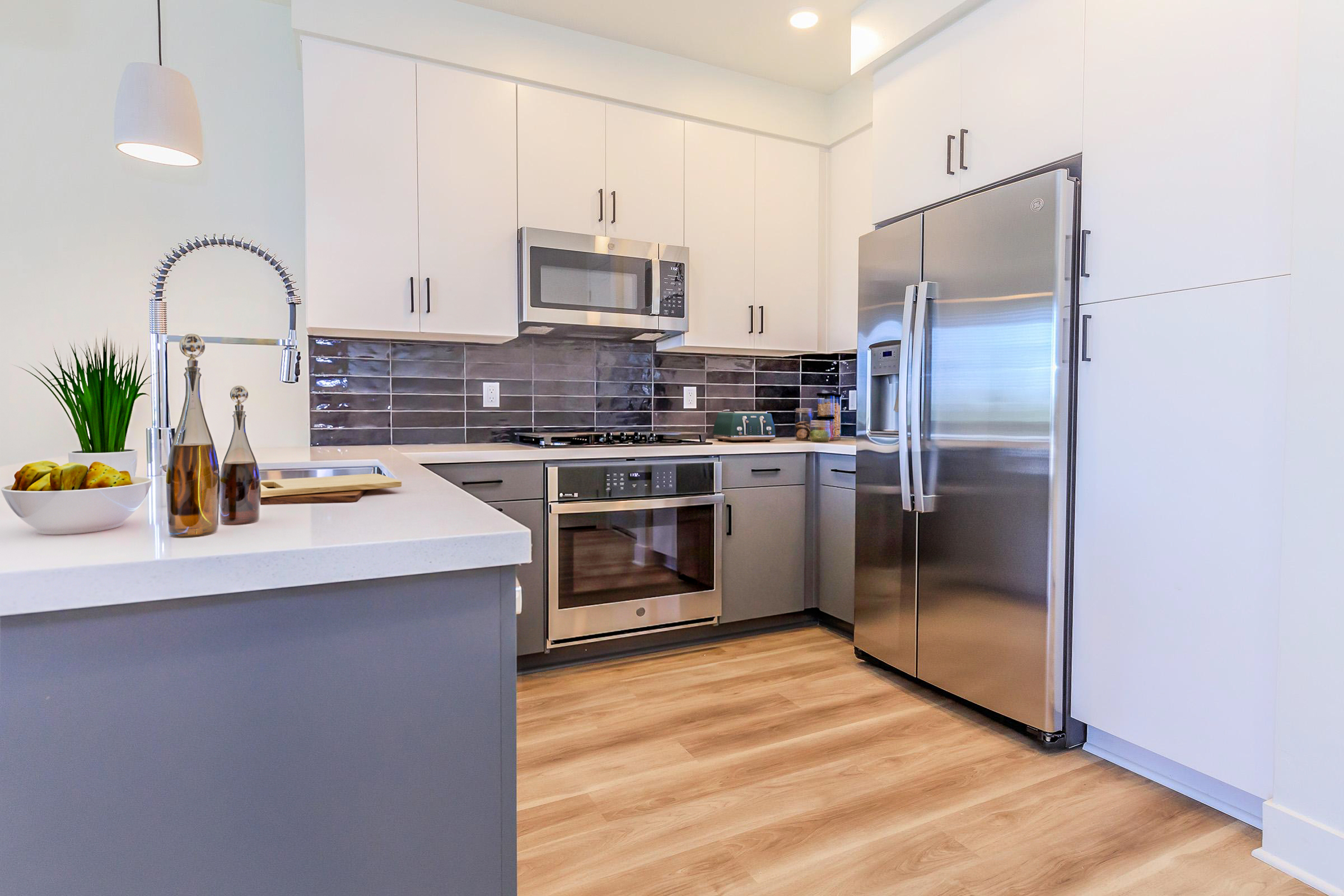 A modern kitchen featuring sleek white cabinetry, stainless steel appliances including a refrigerator and microwave, a gray countertop, and a kitchen island. The floor is made of light wood, and there's a bowl of bananas and glass olive oil bottles on the countertop, along with decorative plants.