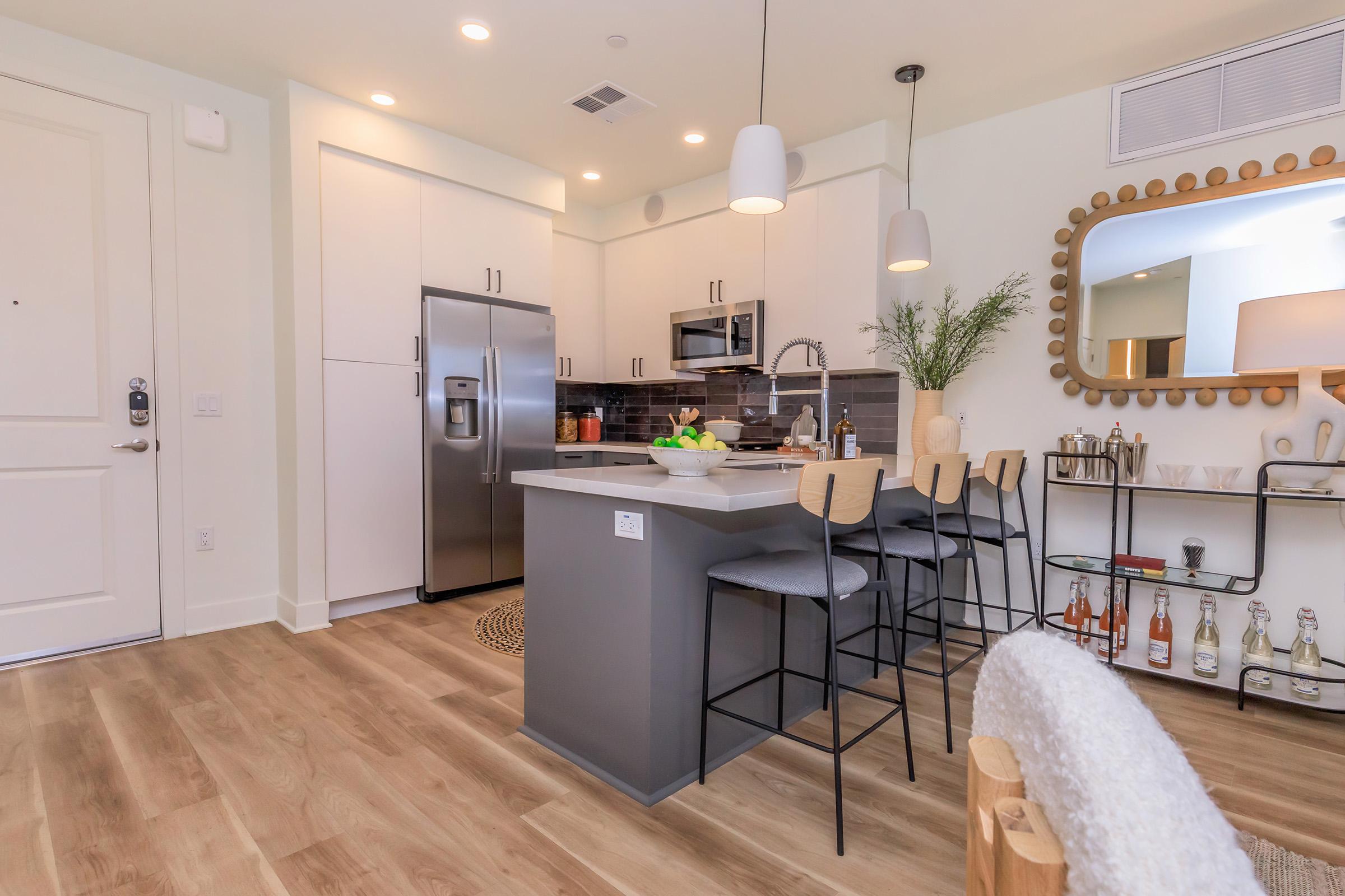 Modern kitchen featuring stainless steel appliances, a large island with seating, and stylish decor. Includes a refrigerator, microwave, and a decorative mirror. The space is well-lit with pendant lighting and has a cozy, inviting atmosphere adorned with plants and tasteful accessories.