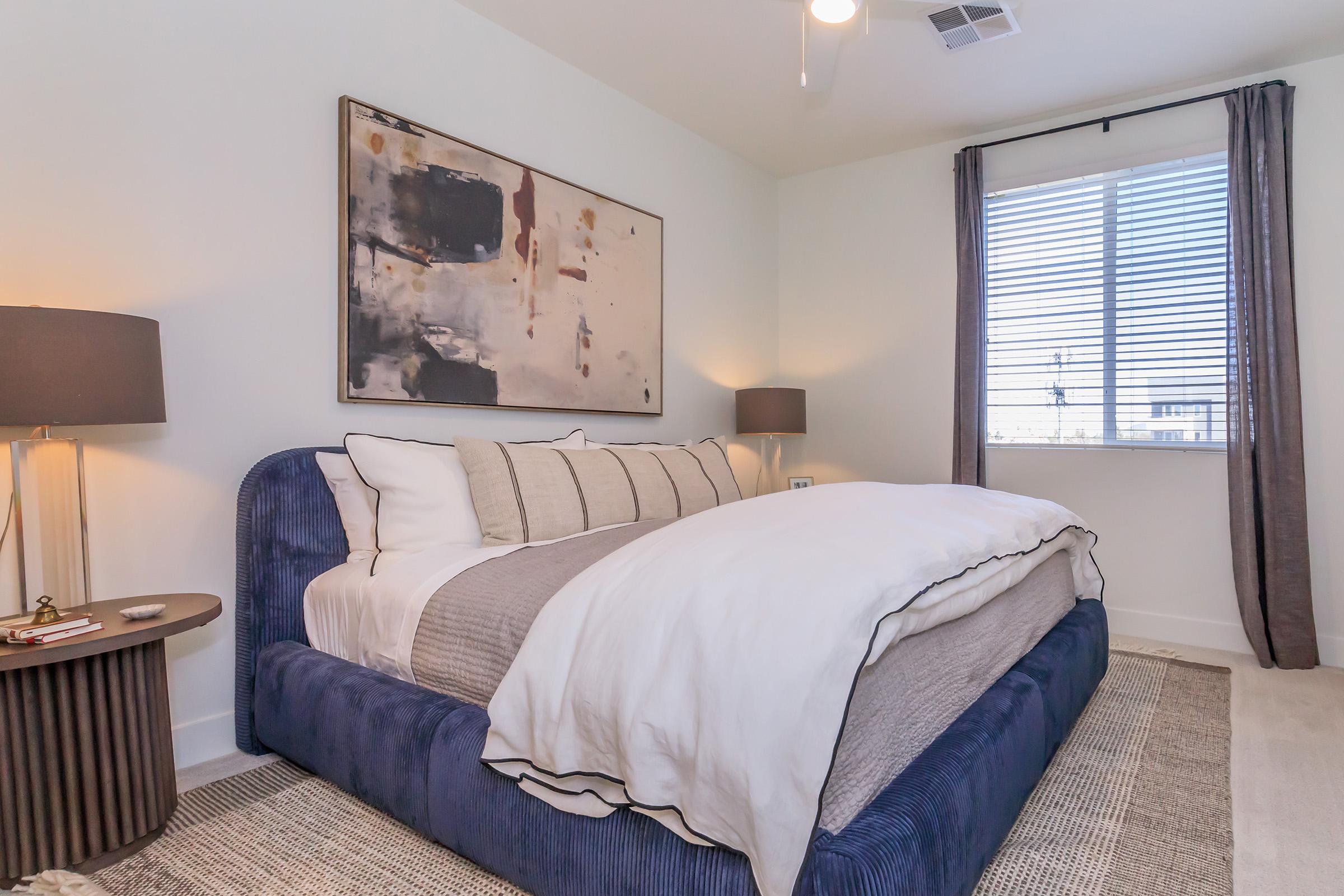 Modern bedroom featuring a blue upholstered bed with beige and white bedding. A stylish area rug covers the floor, and a small round side table with a lamp is beside the bed. Natural light streams through a window adorned with gray curtains. Abstract art hangs on the wall, adding a touch of sophistication.