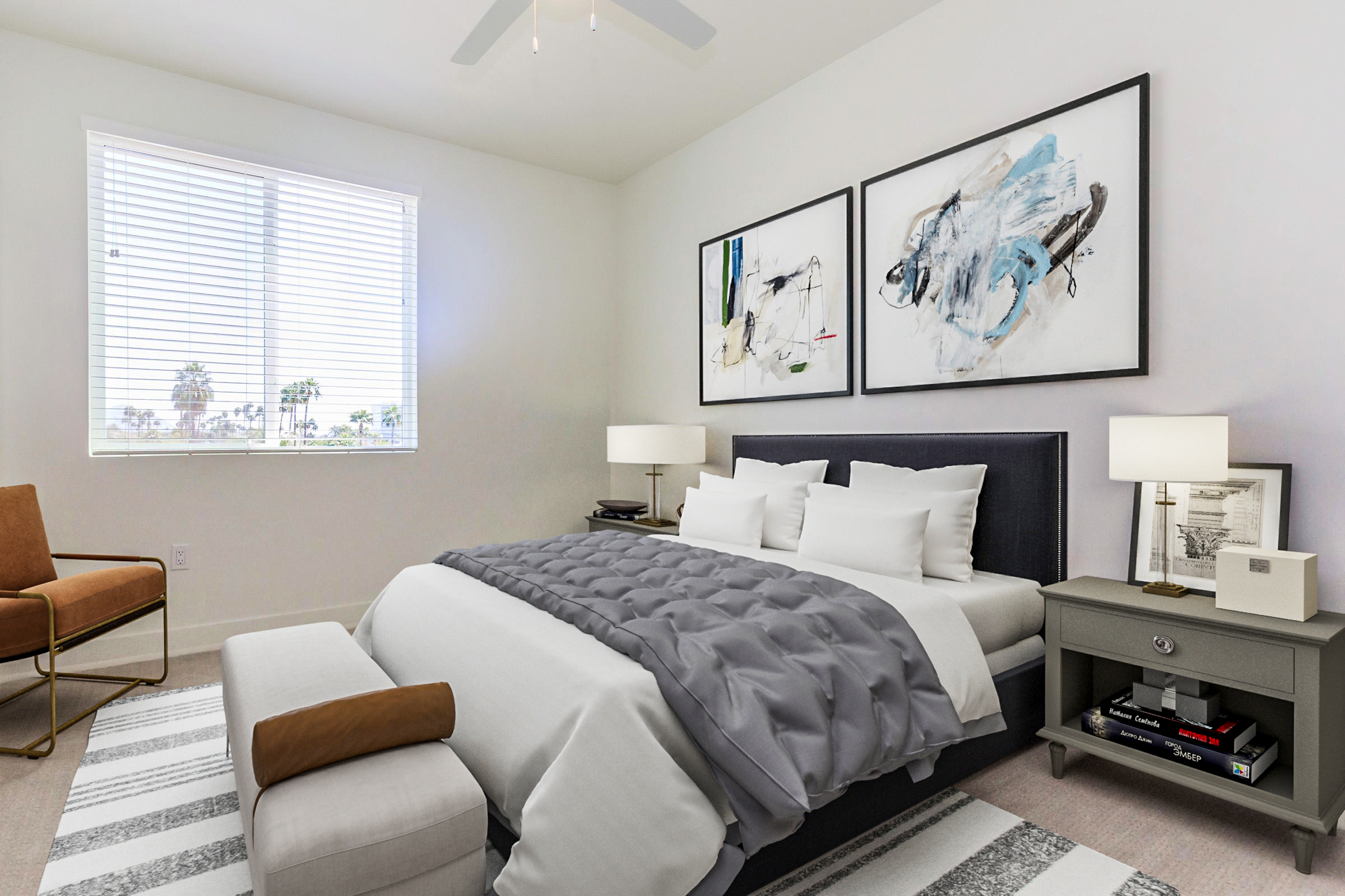A modern bedroom featuring a neatly made bed with gray and white bedding, two abstract paintings on the wall, a nightstand with books and a lamp, a comfortable armchair, and a window with blinds letting in natural light. The room has a minimalistic design with a neutral color palette.