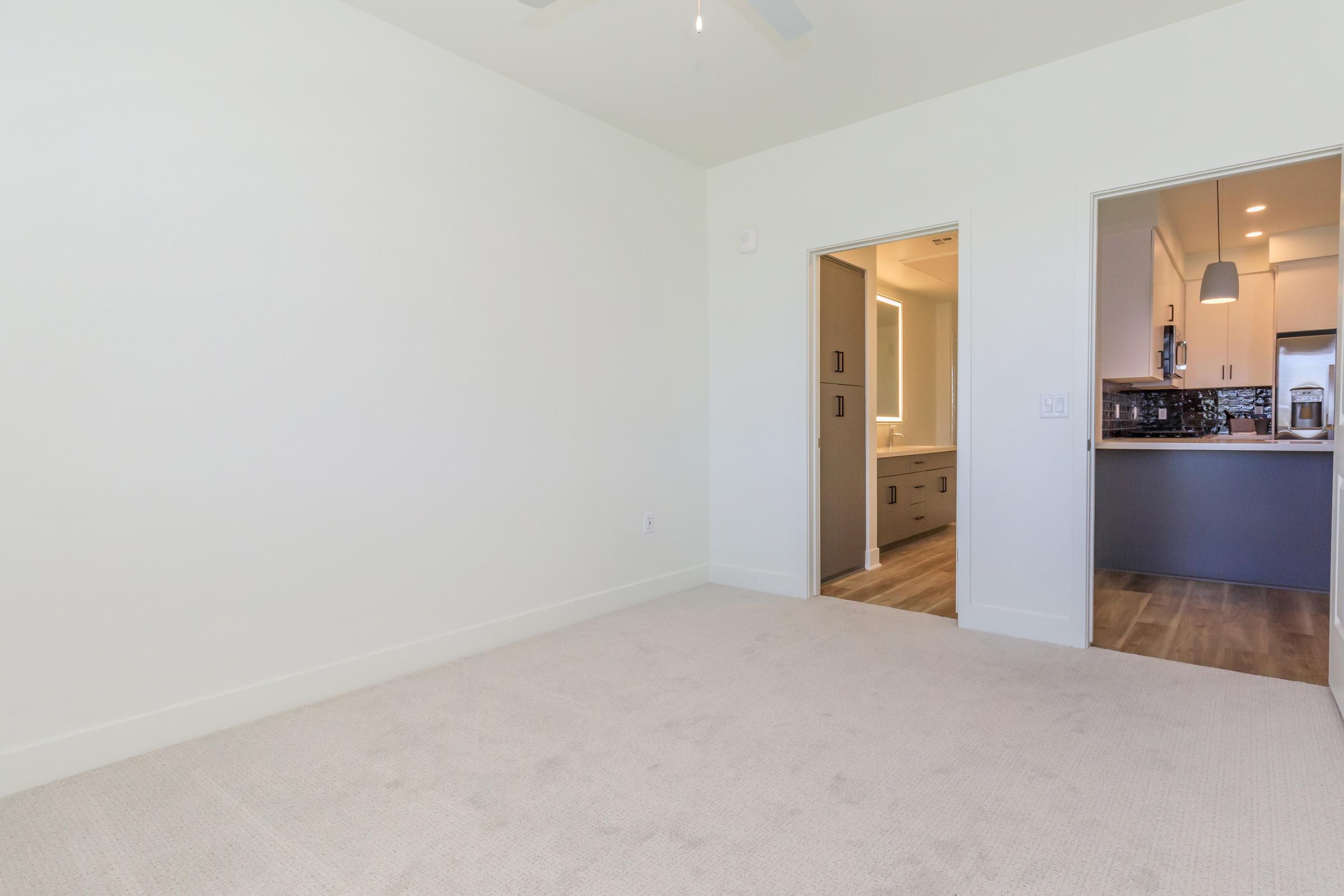 A spacious and minimally decorated room with light-colored walls and carpet. A doorway leads to a kitchen area with modern cabinetry and lighting. The room is well-lit and has a ceiling fan, creating a bright and airy atmosphere.