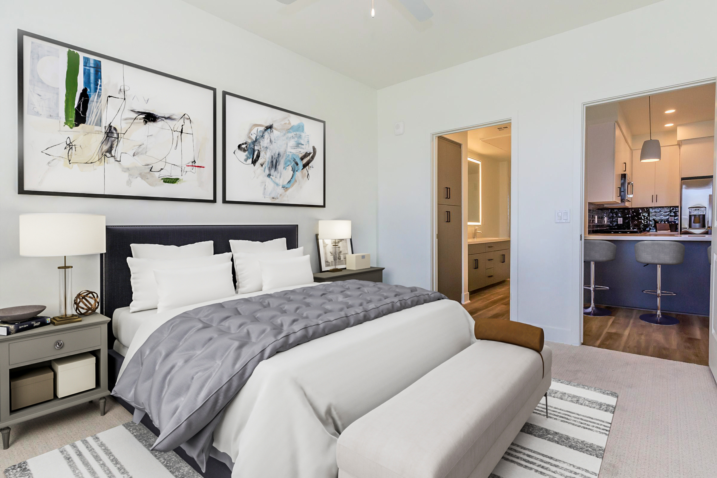 A modern bedroom featuring a large bed with gray and white bedding, two abstract art pieces on the wall, a bedside table with a lamp, and a stylish bench at the foot of the bed. A doorway leads to a kitchenette visible in the background. The overall decor is elegant and contemporary.