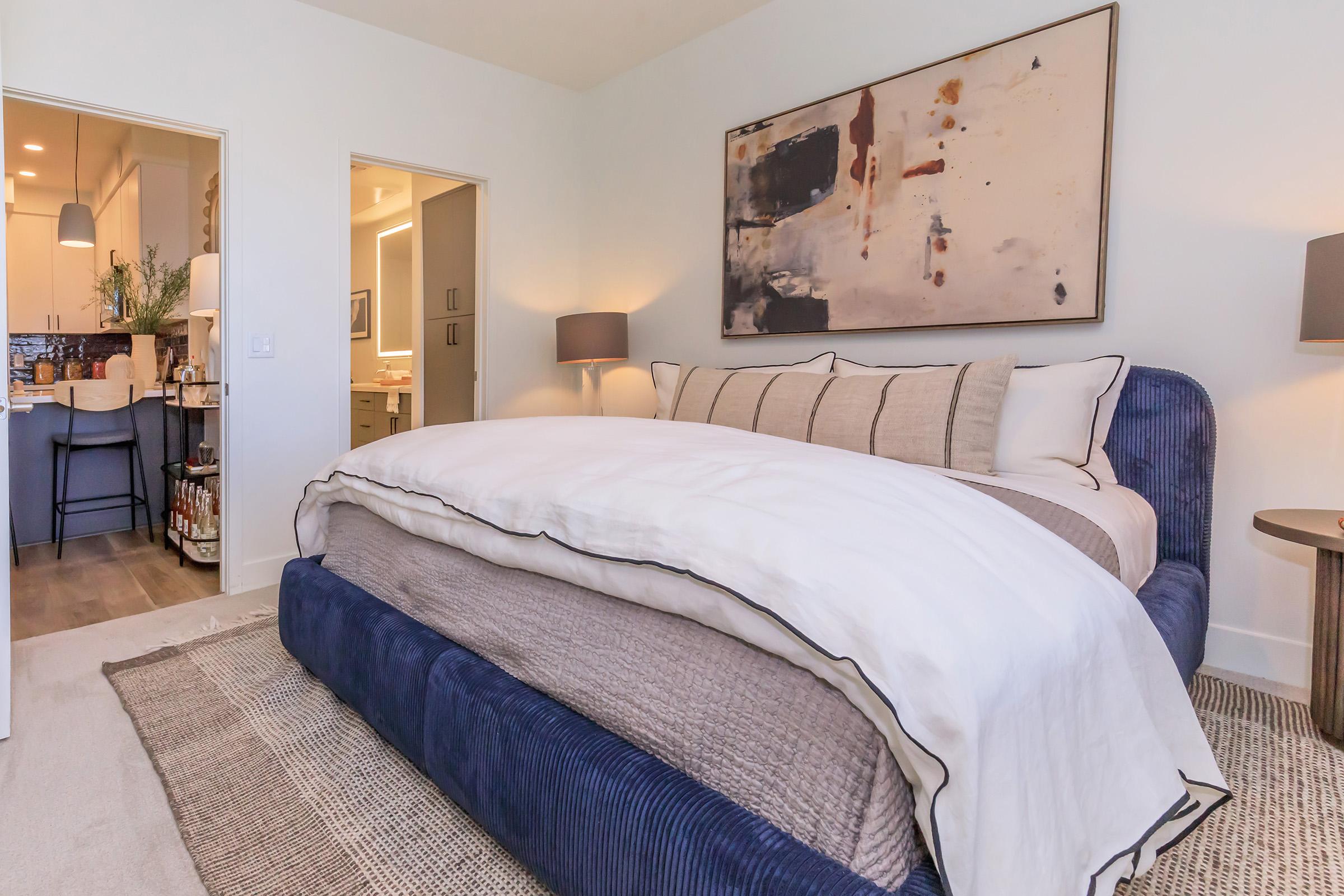 A modern bedroom featuring a large bed with white bedding and a navy blue upholstered base. Light-colored walls complement a colorful abstract painting above the bed. There's a nightstand with a lamp next to the bed, and open doorways lead to other rooms, suggesting a cozy, stylish living space.