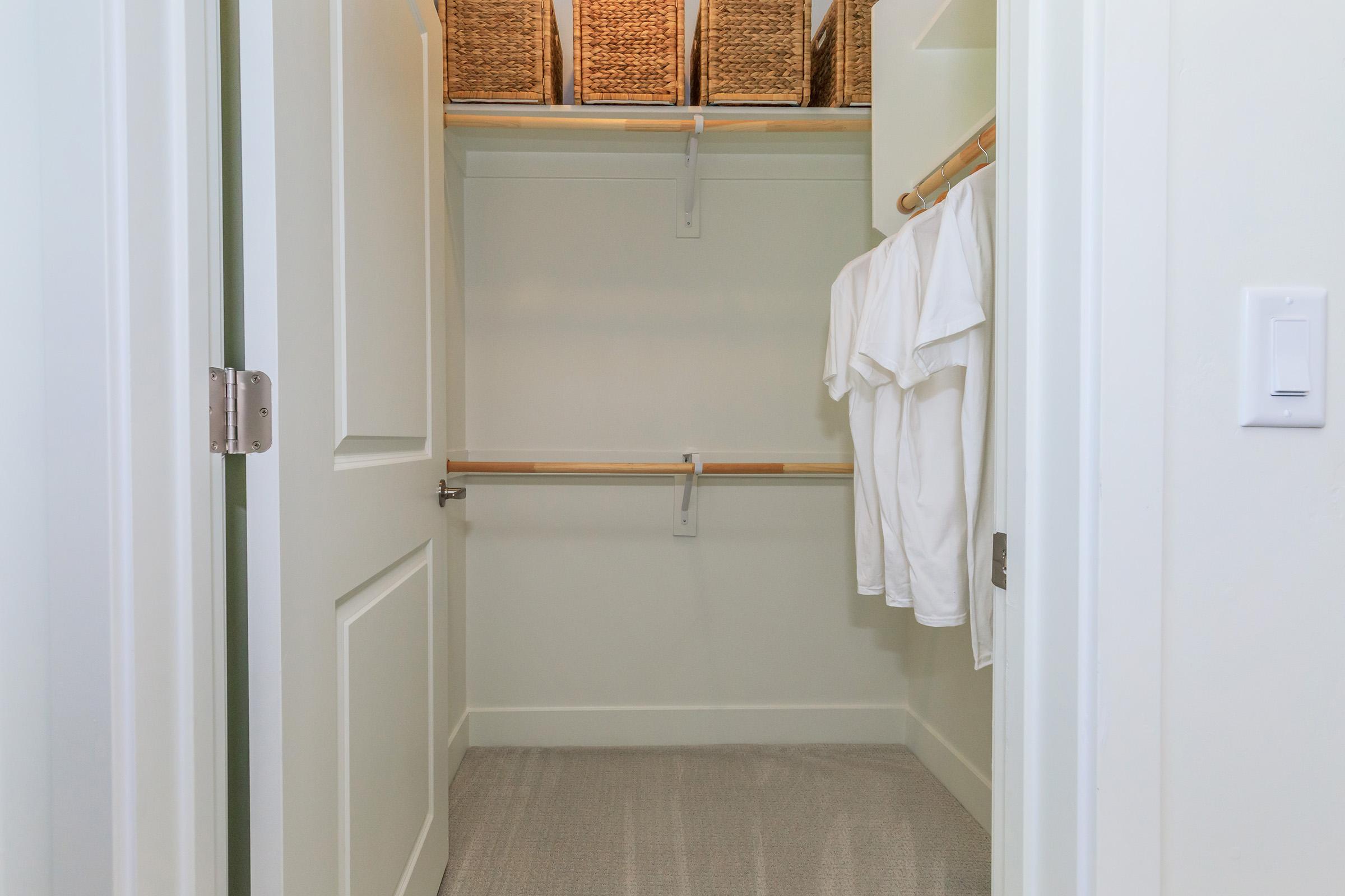 A spacious, organized closet featuring a clean, neutral interior. Two wooden hanging rods hold several white shirts, while woven baskets are neatly arranged on shelves above. The floor is carpeted, and the closet door is open, displaying the uncluttered space.