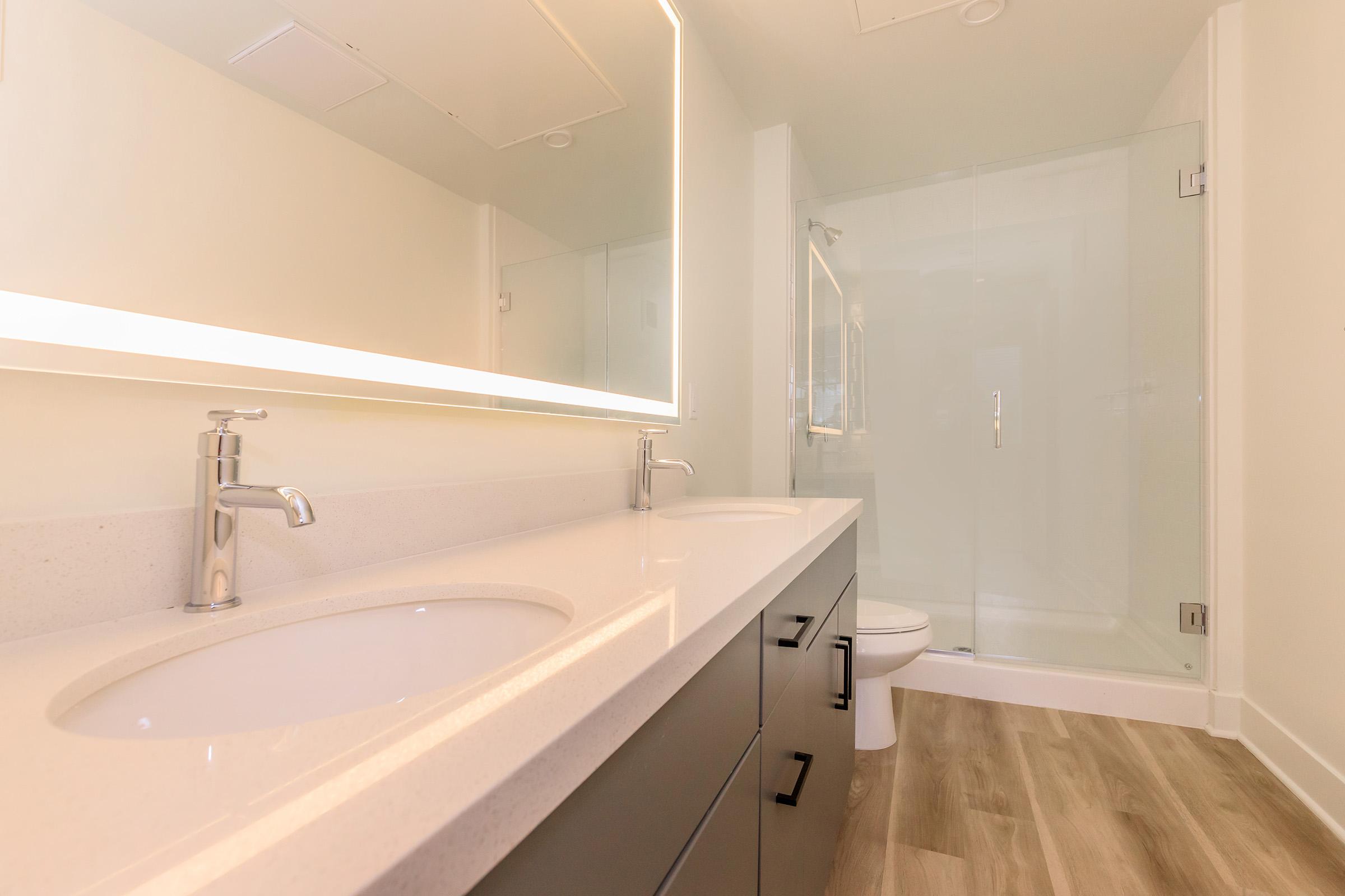 Modern bathroom with a double vanity featuring two sinks, sleek faucets, and a lighted mirror. The space has light-colored walls, a glass shower stall, and wooden flooring, creating a clean and contemporary aesthetic.