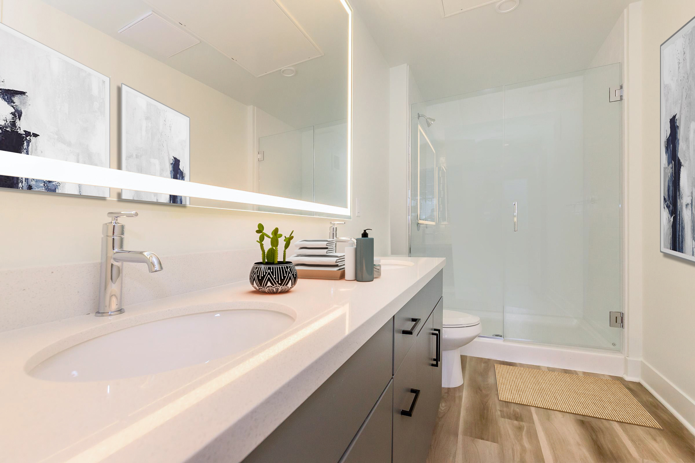 Modern bathroom featuring a double sink vanity with a sleek countertop, large mirror, and contemporary faucets. A small plant decorates the space, alongside neatly stacked towels. The walk-in shower is enclosed with glass, and the flooring has a wood-like finish, creating a bright and stylish atmosphere.