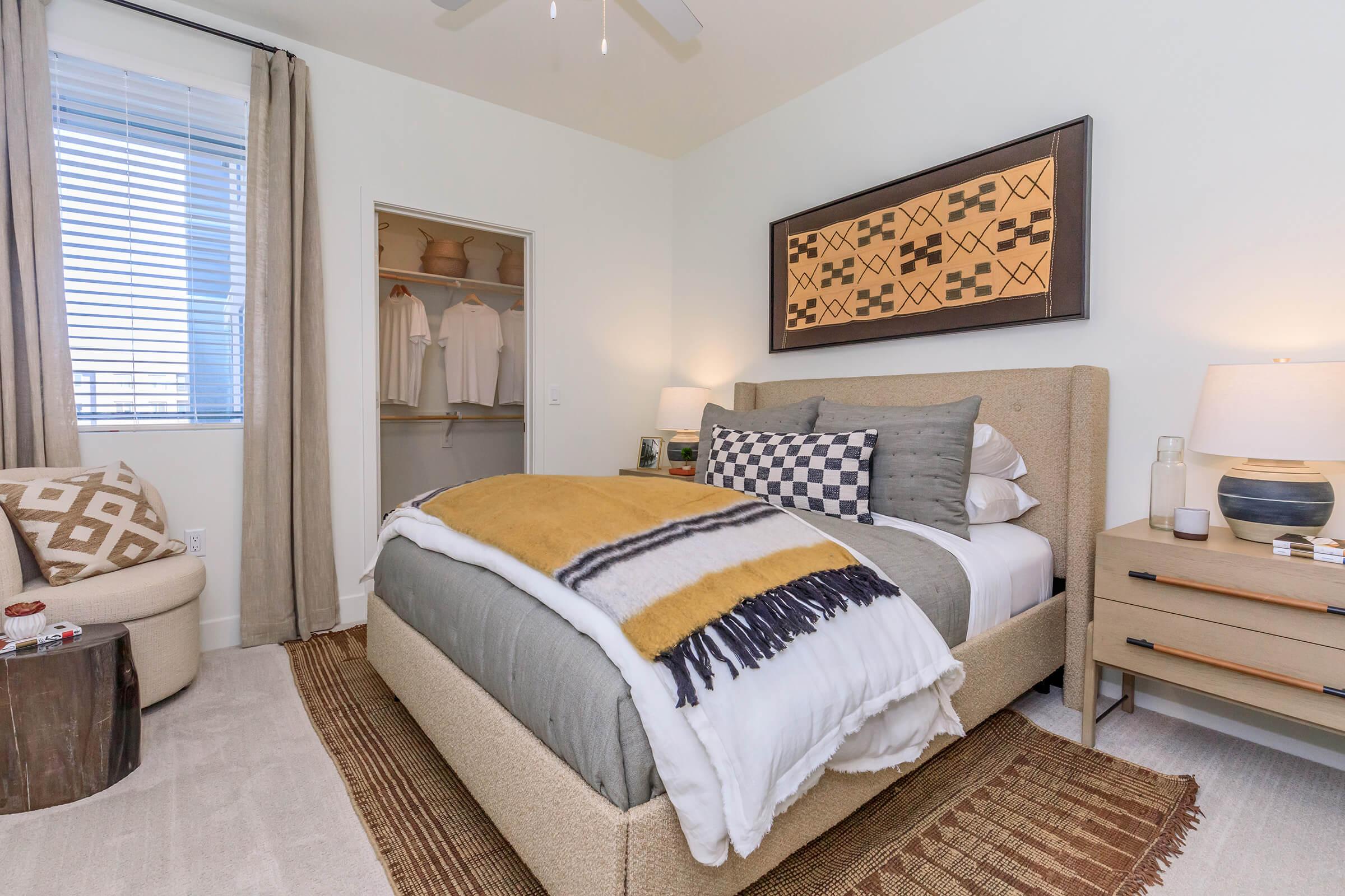 A cozy bedroom featuring a beige bed with layered blankets, decorative pillows, and a patterned throw. A bedside table holds a lamp, and a woven rug covers the floor. Natural light streams through a window with light curtains. A closet is partially visible, showcasing clothing items. Décor includes artwork on the wall and a textured chair.