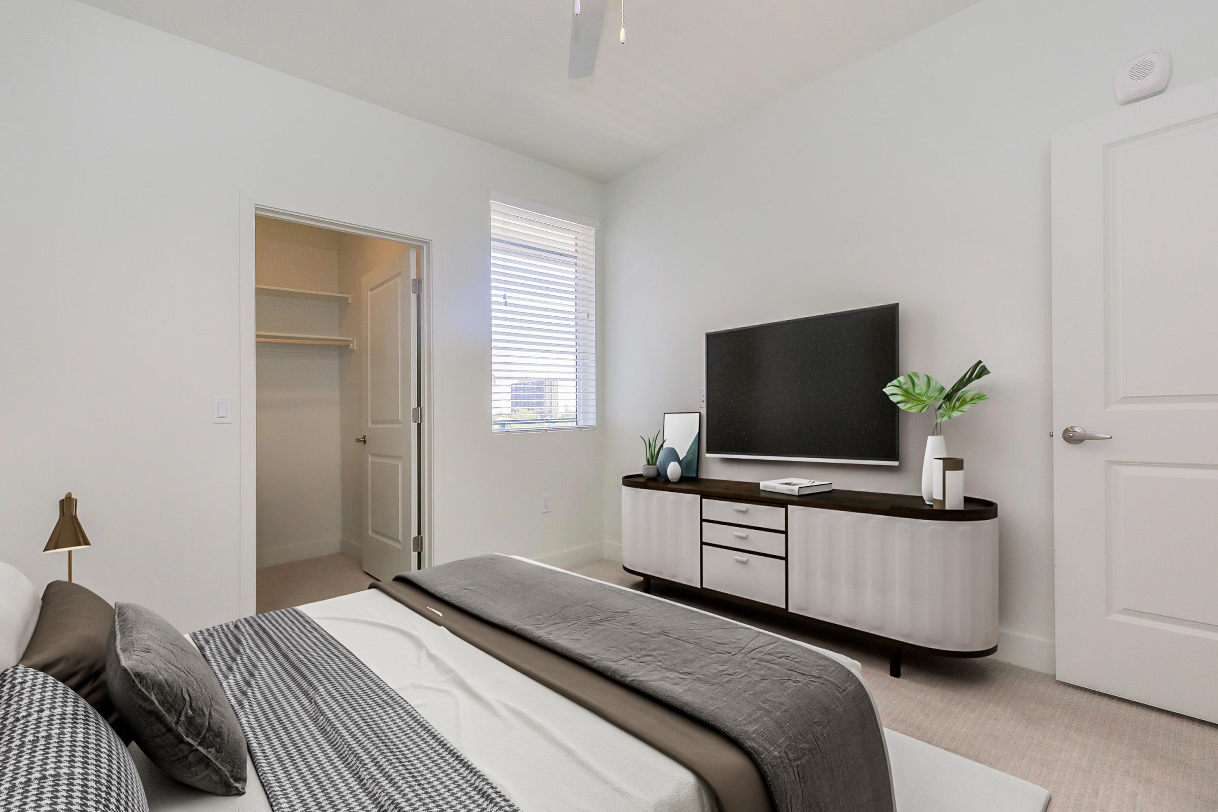 A modern bedroom featuring a neatly made bed with gray and white bedding, a sleek dresser with a television on top, a potted plant, and a window letting in natural light. There's a closet door visible in the background, and the walls are painted in a light color, creating a spacious and inviting atmosphere.