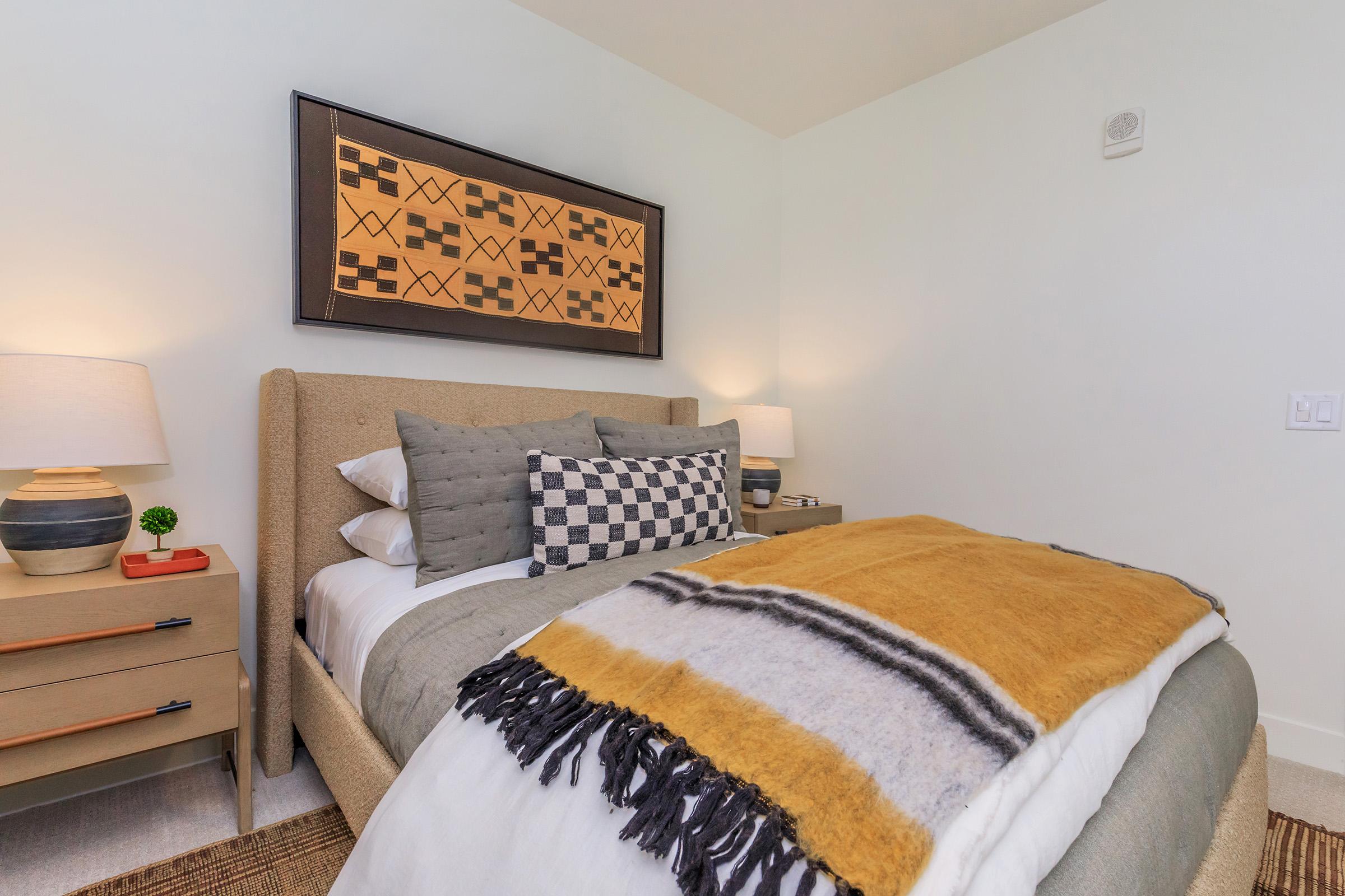 Cozy bedroom featuring a neutral palette with a bed adorned with decorative pillows and an orange and gray throw blanket. Two nightstands with lamps flank the bed, and a patterned artwork hangs above the headboard, adding a touch of style to the serene space.