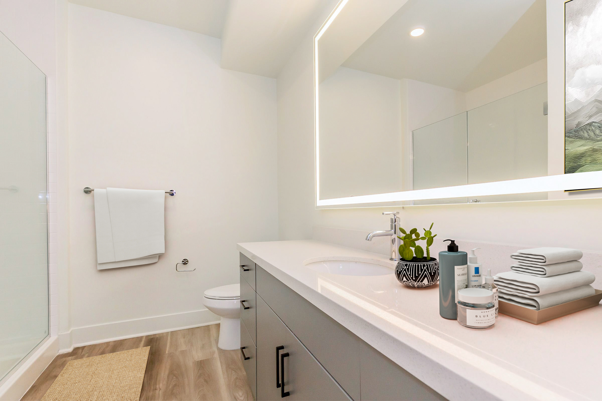 A modern bathroom featuring a sleek countertop with toiletries, neatly arranged towels, and a small potted plant. The space is bright, with a large mirror illuminated by soft lighting, and a shower area visible in the background. The decor is clean and minimalist, creating a serene atmosphere.
