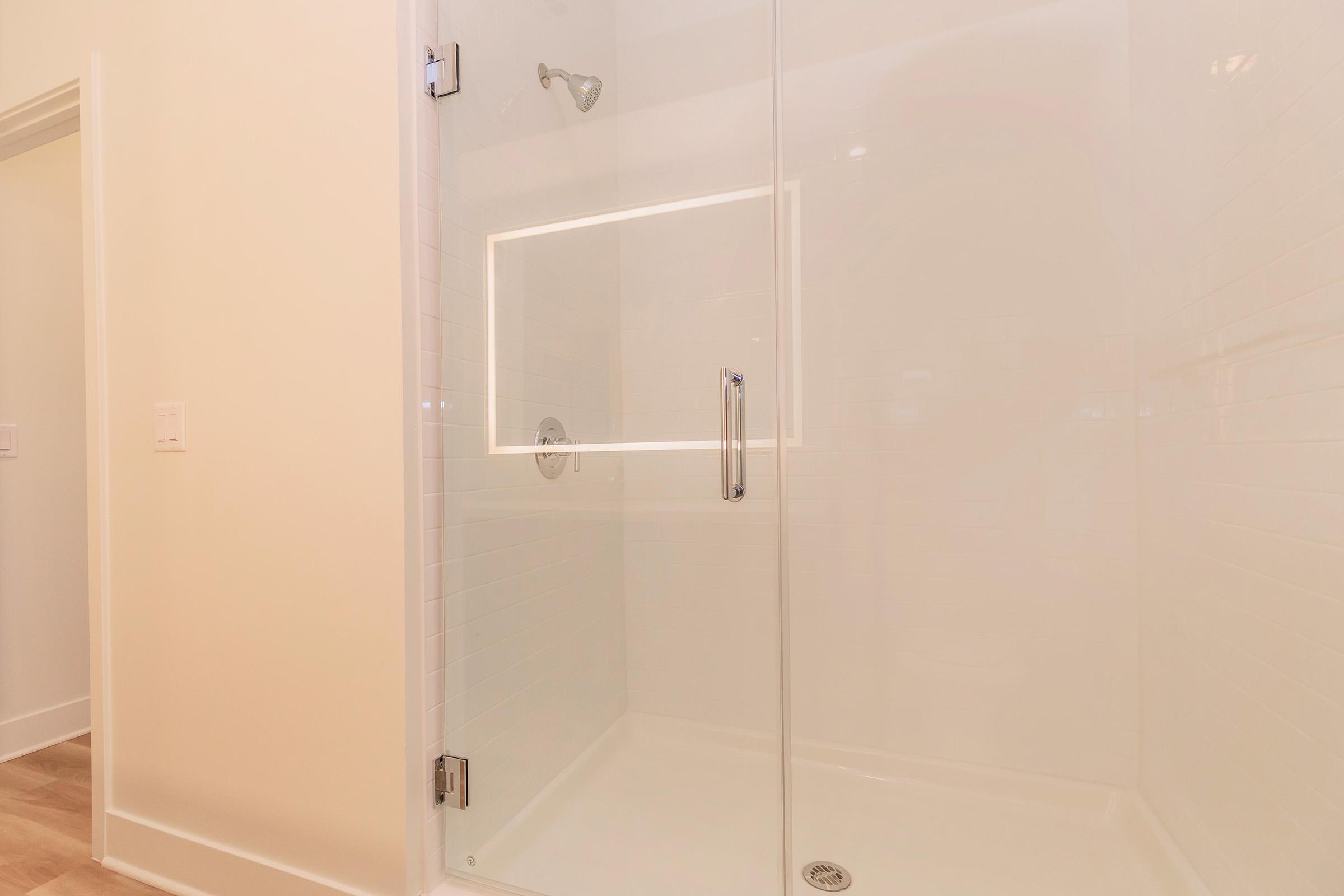 A modern shower with a glass enclosure and a chrome showerhead. The walls are lined with white tiles, and there is a sleek, minimalist design. The floor is hardwood, and there is a clear view of the spacious shower area, enhancing the bright and airy feel of the bathroom.