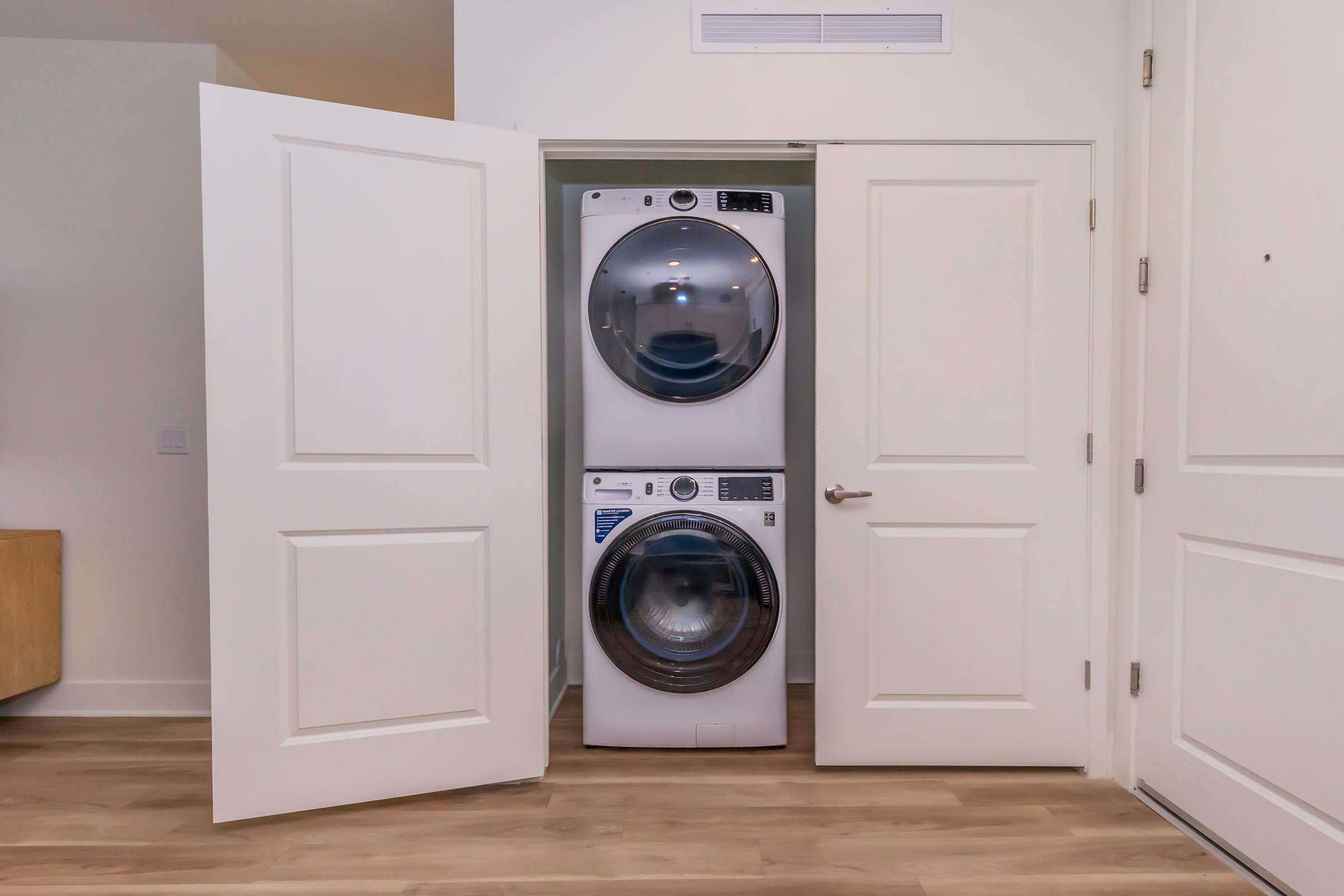 A stacked washer and dryer unit neatly tucked between two open white doors in a modern interior space. The flooring is wood-look, and the walls are clean and light-colored, contributing to a minimalist design.