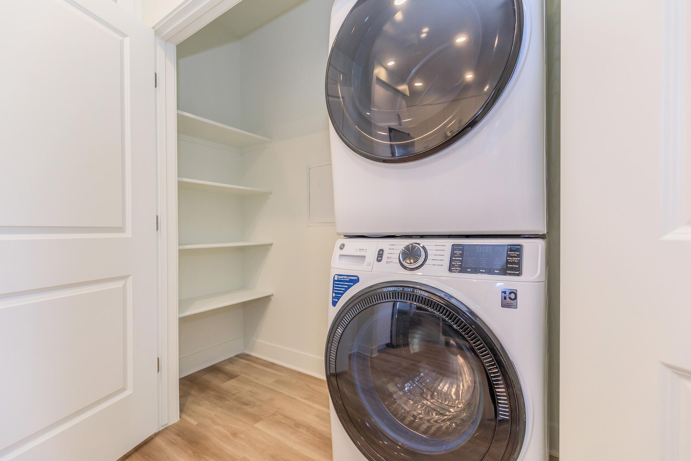 A modern laundry room featuring a stacked washer and dryer set in a clean, minimalistic space. The walls are painted a soft color, and open shelves are visible to the left. The floor is light wood, creating a bright and airy atmosphere.
