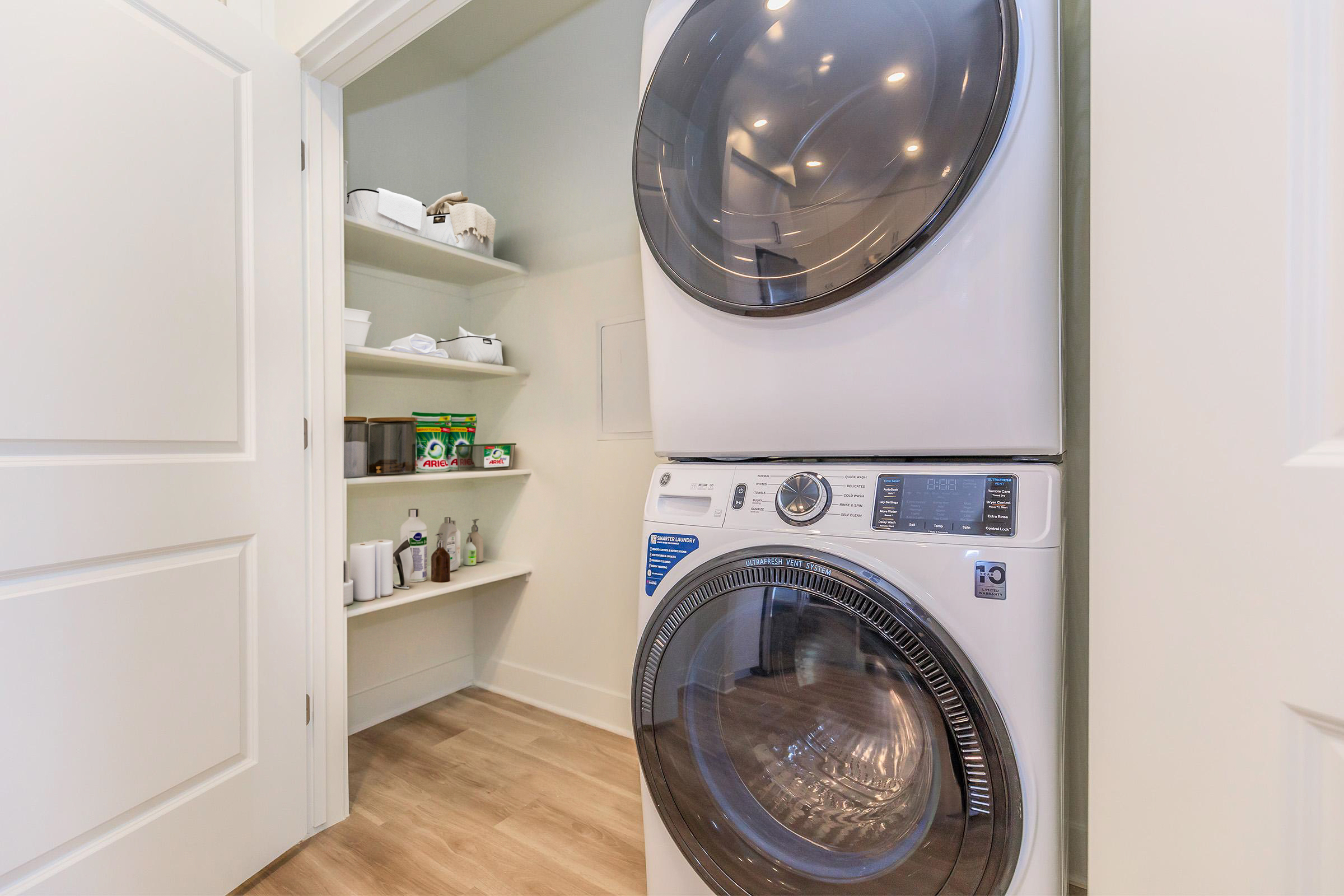 A laundry area featuring a stacked washer and dryer. The space includes a shelf visible in the background with various containers and cleaning supplies. The room has light-colored walls and wooden flooring, providing a tidy and organized appearance.