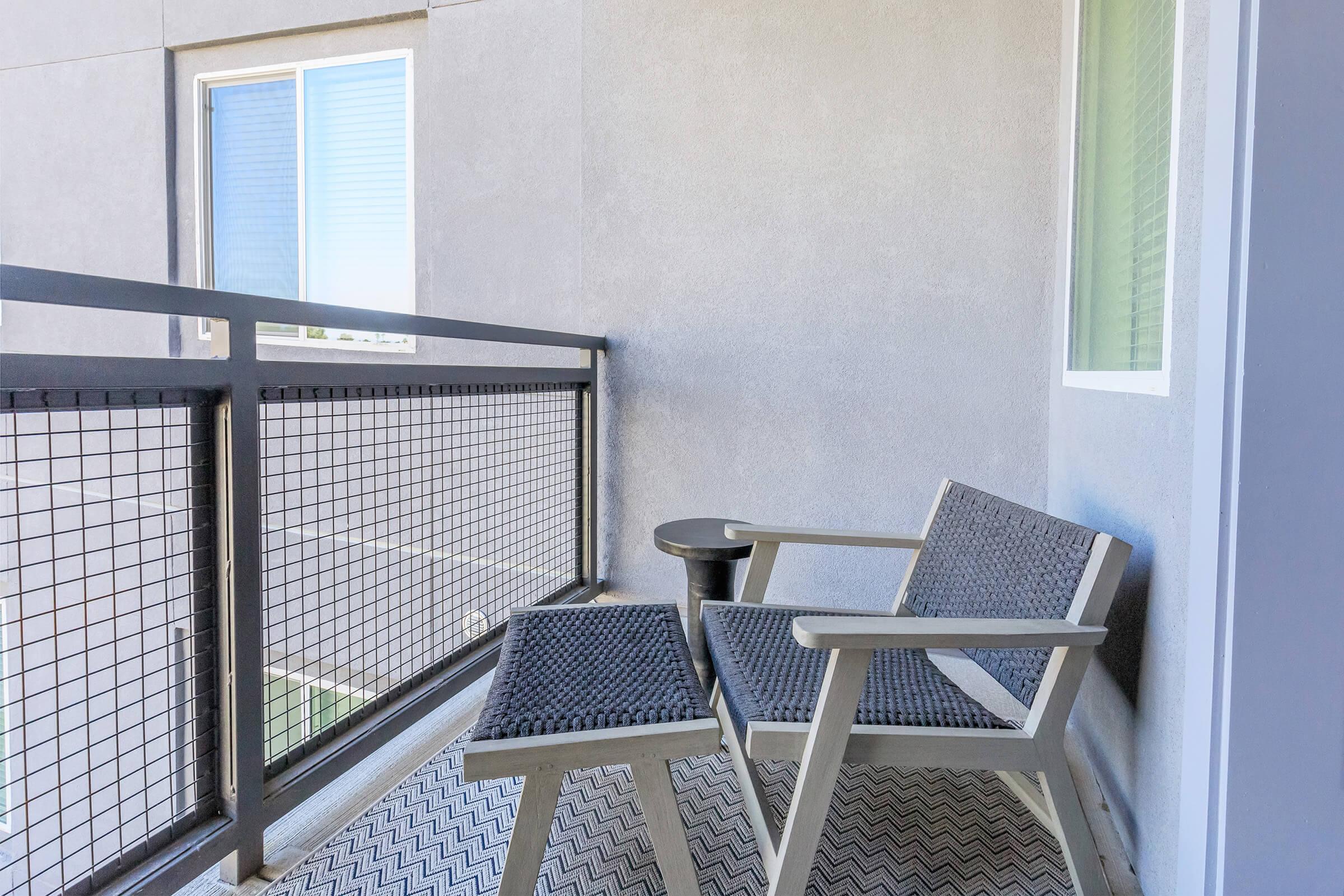 A small balcony with two gray wicker chairs and a round black table, overlooking a light-colored wall and a window. The flooring features a herringbone pattern, and there is natural light entering through the nearby window.