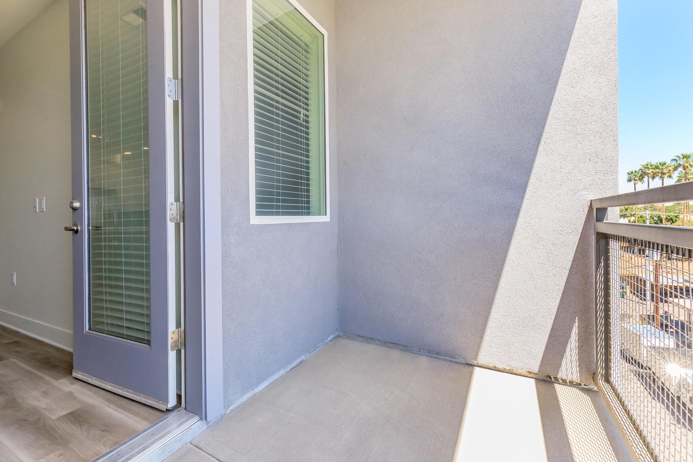 A small balcony with a concrete floor and a glass door leading inside. The wall is smooth and gray, while a window with horizontal blinds is visible. Sunlight casts shadows on the floor, and palm trees can be seen in the distance beyond the railing.