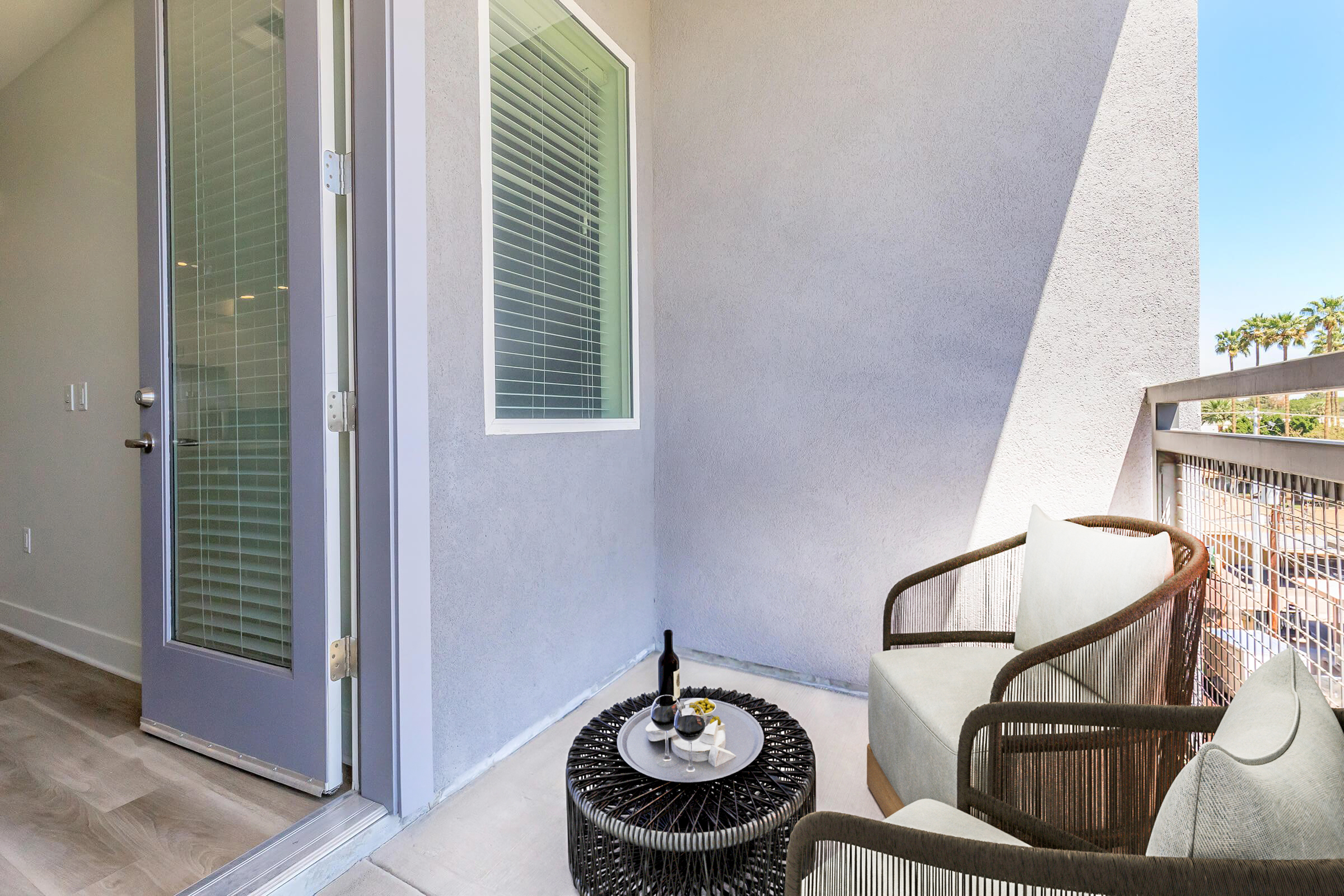 A modern balcony featuring a round table with a bottle of wine and snacks, flanked by two stylish chairs. The wall is painted in a light gray, and there is a sliding glass door leading inside. Sunlight brightens the space, with a glimpse of palm trees visible in the background.