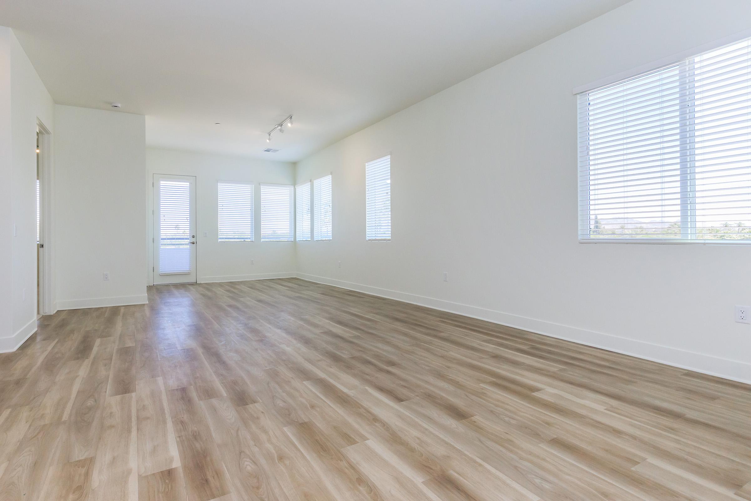 Spacious, empty room with large windows featuring blinds, hardwood-like flooring, and white walls. Natural light fills the space, creating a bright and open atmosphere. Ideal for various interior design choices.