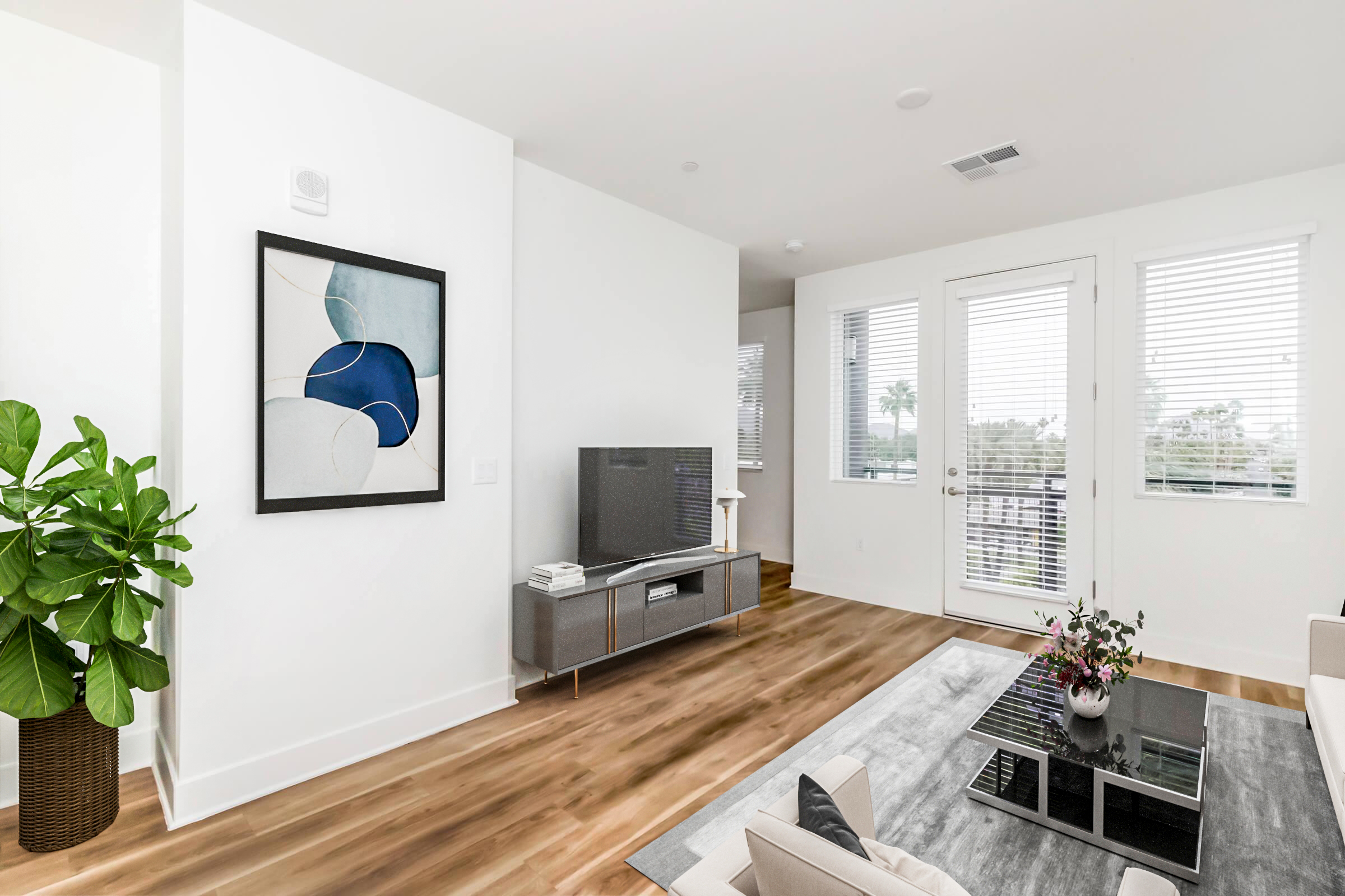 A modern living room featuring a white wall with abstract artwork, a television on a sleek gray media unit, and a coffee table with a small vase of flowers. Large windows with blinds allow natural light into the space, and greenery is present in a potted plant near the wall, enhancing the fresh and airy atmosphere.
