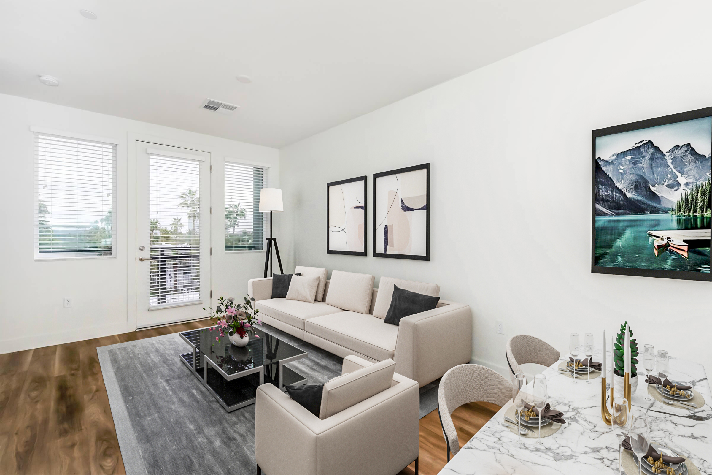 Modern interior of a bright living room featuring a light-colored sofa, a glass coffee table, and a dining area with a marble tabletop. The walls are adorned with framed artwork, and large windows allow natural light to fill the space. A lamp stands beside the sofa, and plants add a touch of greenery.