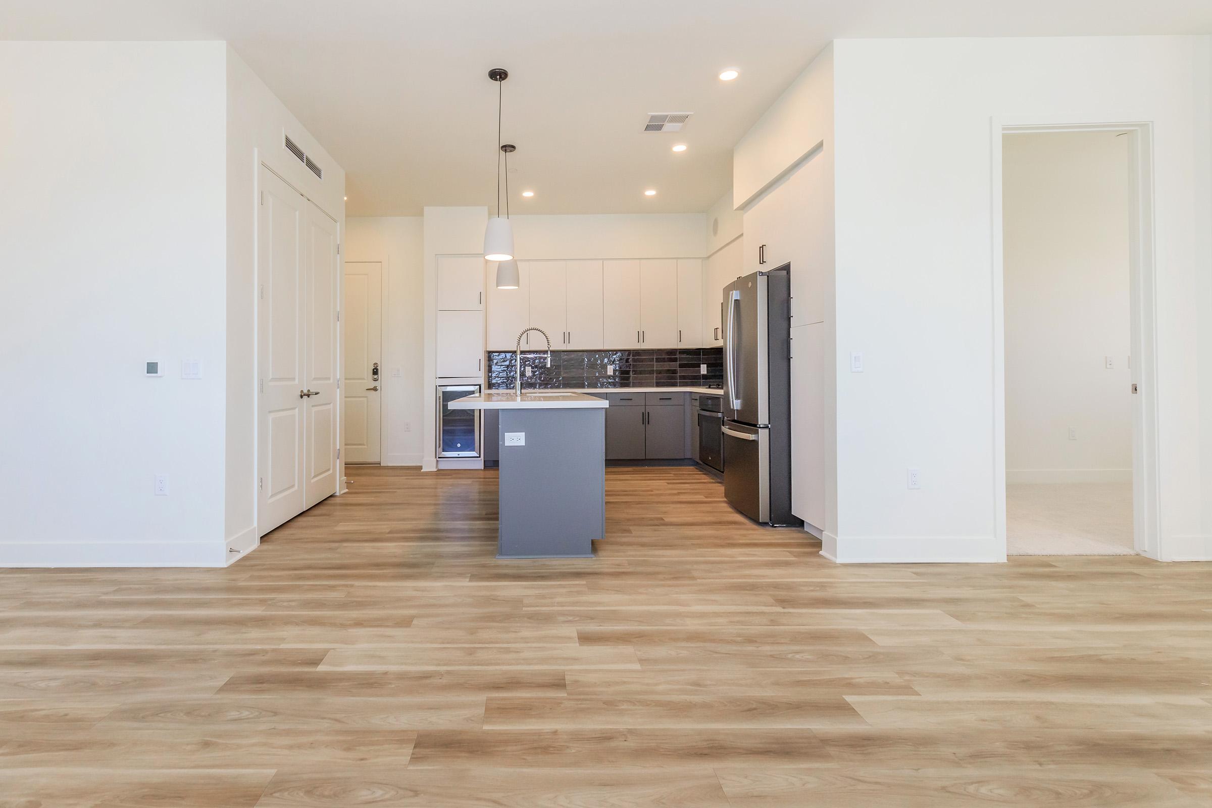 Modern kitchen and living area with light wood flooring. Features include a central island with a sink, stainless steel appliances, and minimalist cabinetry. Bright, open layout with large windows allowing natural light. White walls and sleek design create a contemporary feel.