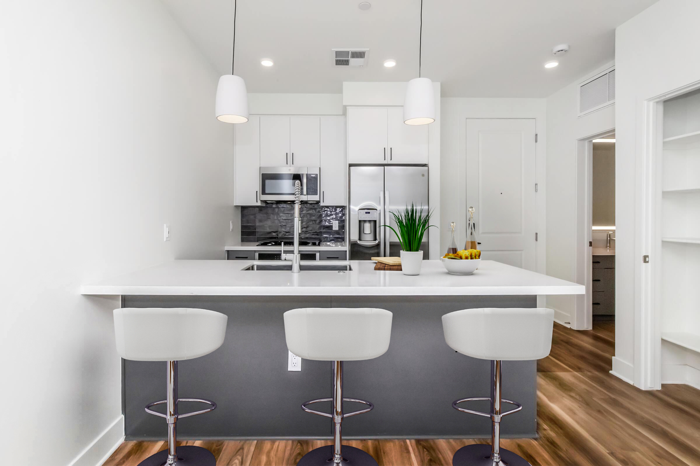 Modern kitchen featuring a sleek island with three white bar stools. The countertop is white, complemented by stainless steel appliances and dark cabinetry. Natural light illuminates the space, showcasing a minimalist design with potted plants and a bowl of fruit on the island. A doorway leads to another room.