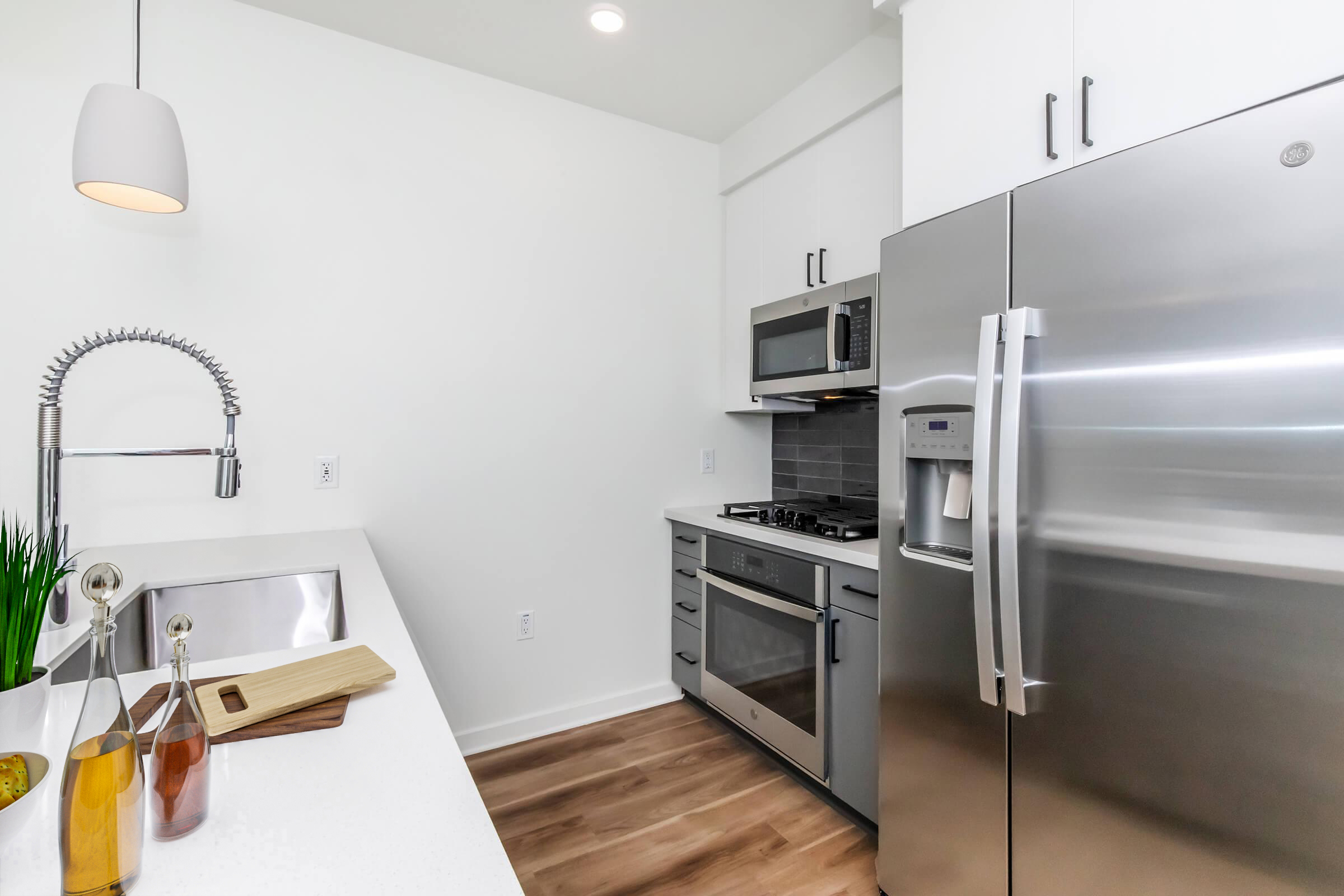 Modern kitchen featuring stainless steel appliances, including a refrigerator, oven, and microwave. A sleek sink with a high-arc faucet is positioned next to a wooden cutting board and decorative bottles. The walls are painted white, and the flooring is light wood, creating a bright and inviting atmosphere.