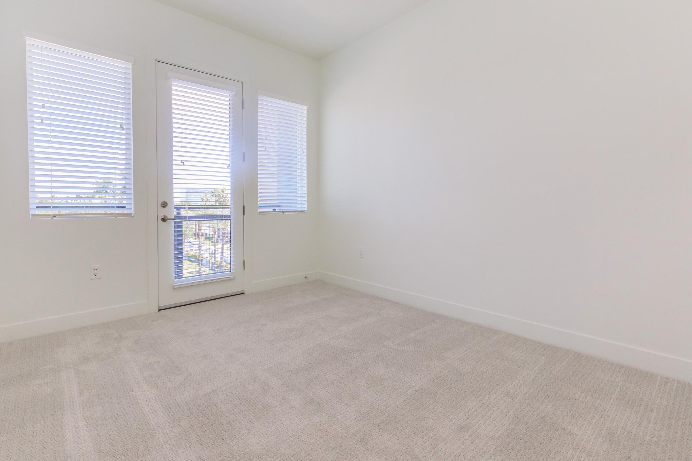 Empty room with light-colored walls and beige carpet. Two windows with white blinds allow natural light, and a glass door leads outside. The room has a minimalist design and appears spacious, offering a serene and uncluttered atmosphere.