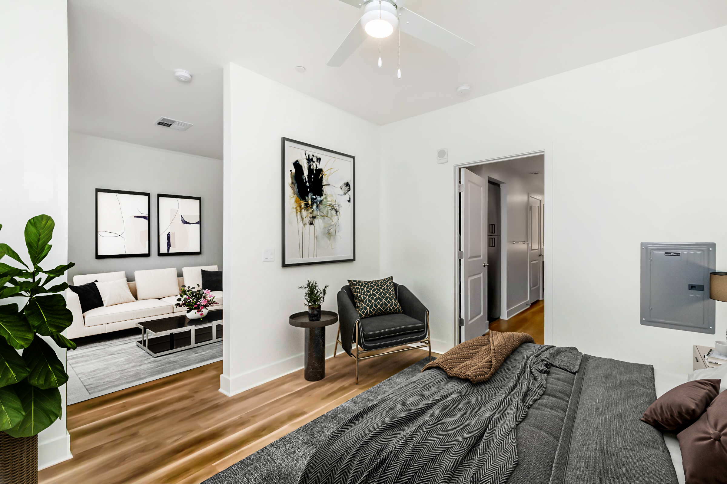 A modern, well-lit bedroom featuring a large bed with dark bedding. A nearby bedside table holds a lamp. In the background, a spacious living area with a beige sofa, a floral arrangement, and artwork on the walls is visible. There’s a plant in the corner and light-colored wooden flooring.