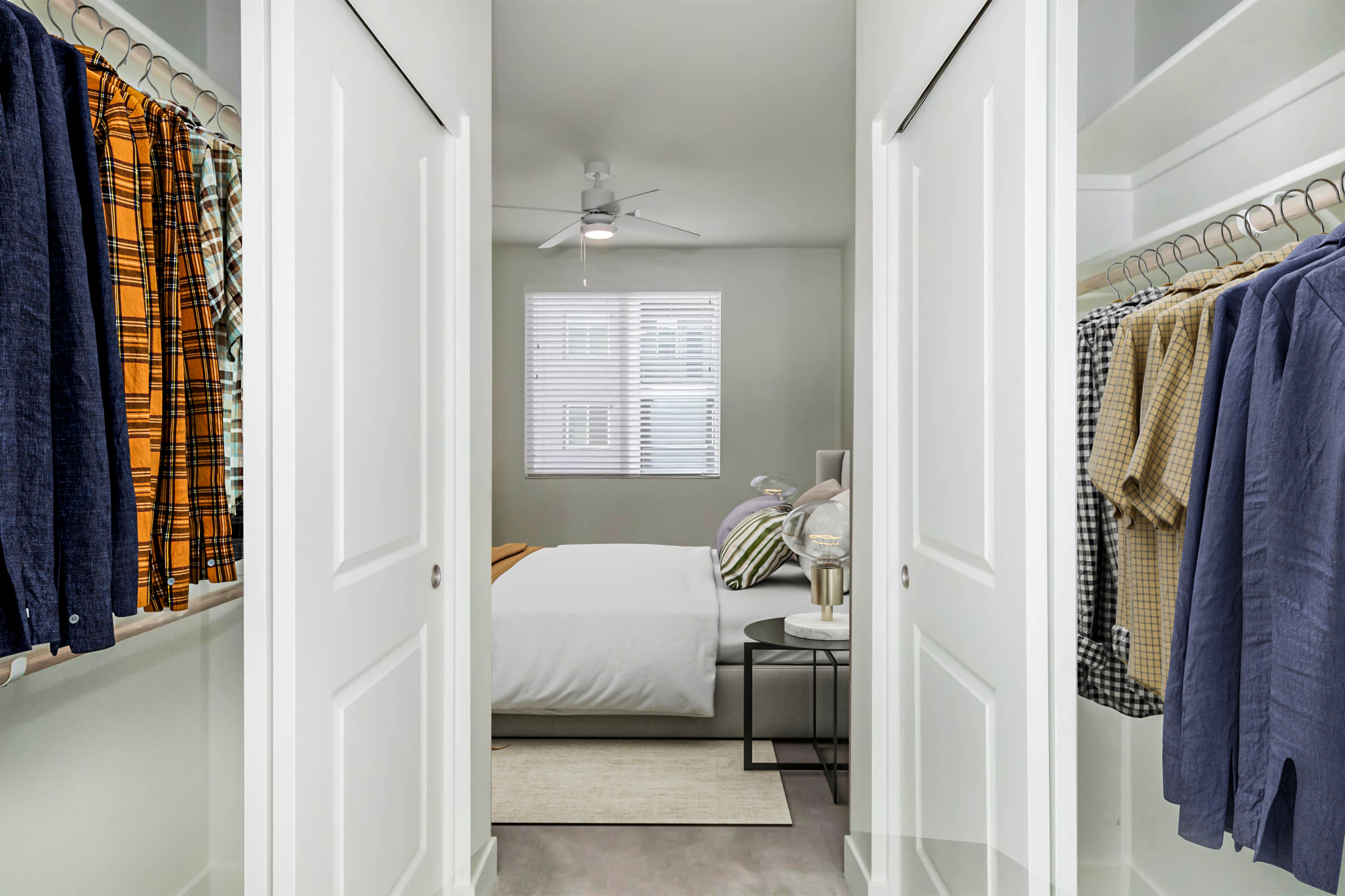 A view from a hallway into a bright bedroom, featuring a white bed, a ceiling fan, and a window with blinds. On either side of the hallway, there are organized closets with clothes hanging on hangers, showcasing a mix of patterns and colors. A small side table is placed beside the bed.