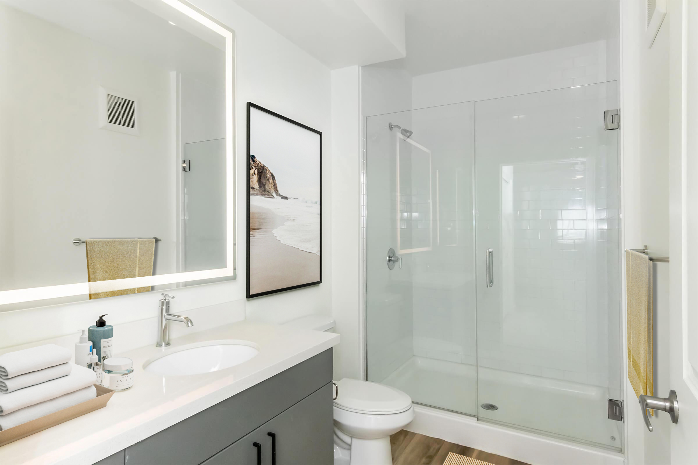 Modern bathroom featuring a glass-enclosed shower, a sleek vanity with a round mirror, neatly arranged towels, and a piece of wall art depicting a beach scene. The color scheme is neutral with light gray cabinetry and wooden flooring, creating a clean and contemporary look.
