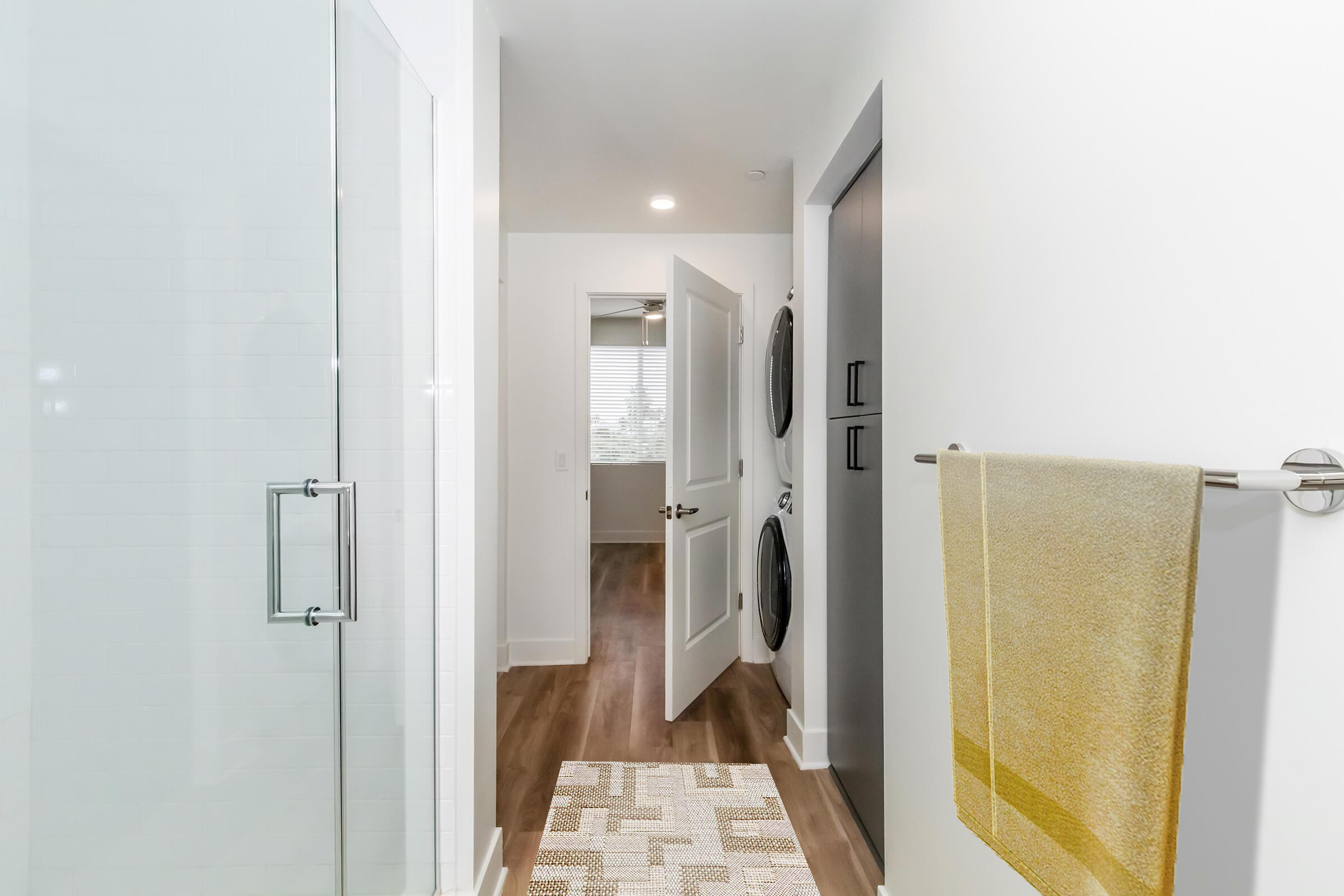 A modern bathroom featuring a walk-in shower with a glass door, a towel hanging on a rack, and a laundry area with stacked appliances visible. The space has light-colored walls, warm wooden flooring, and natural light coming through a window in the distance.