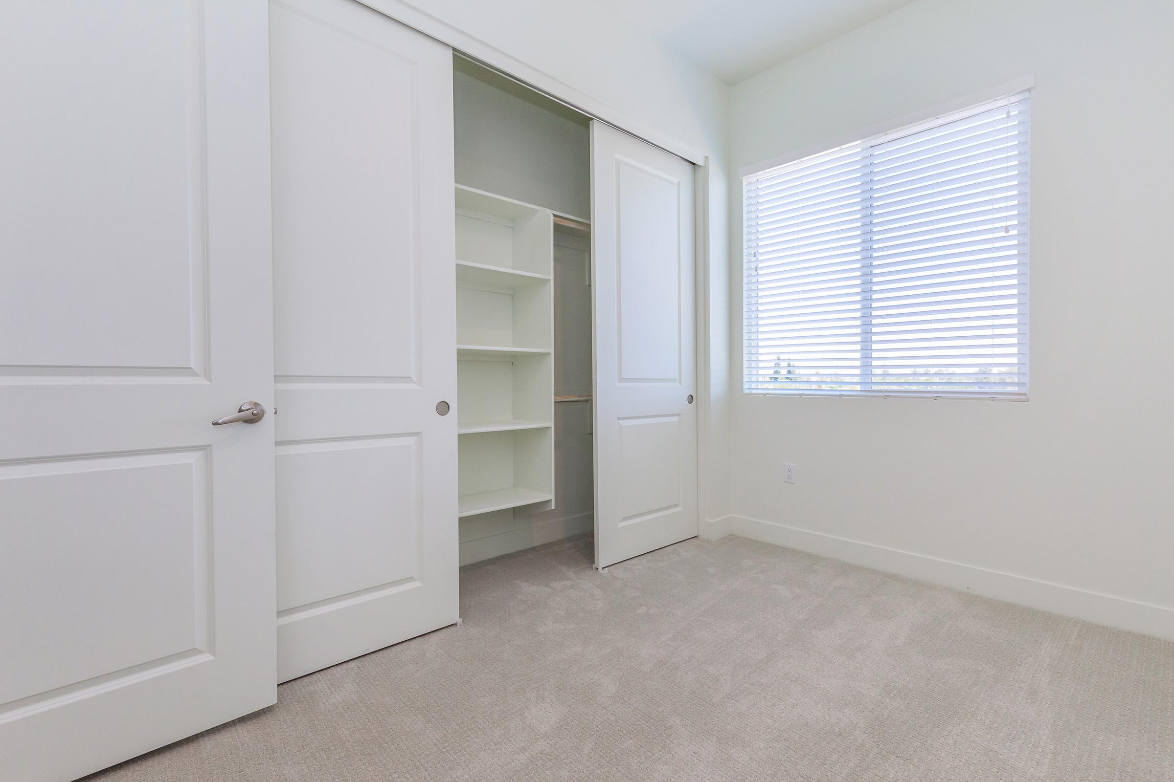 A light-colored room with beige carpet, featuring a large window with blinds on the right side. There is a closet with sliding doors on the left, partially open, revealing shelves inside. The walls are painted in a soft, neutral tone, creating a bright and airy atmosphere.