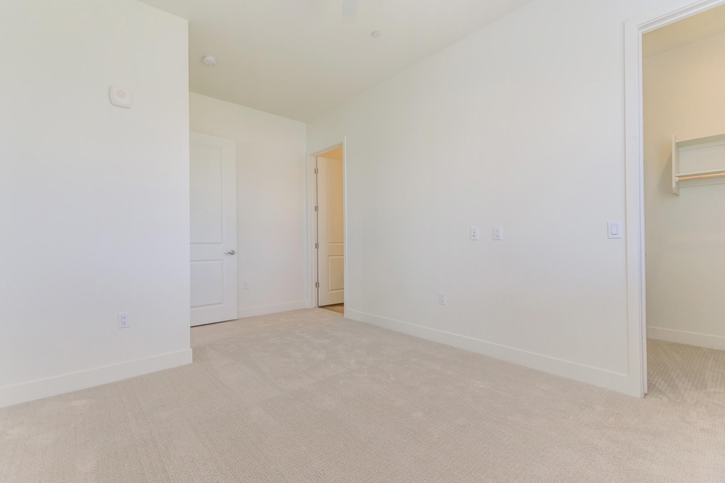 A spacious, empty room with light-colored walls and a beige carpet. There is a white door leading to another room and an open closet with shelving. The overall ambiance is bright and minimalistic, suitable for various uses such as a bedroom or office.