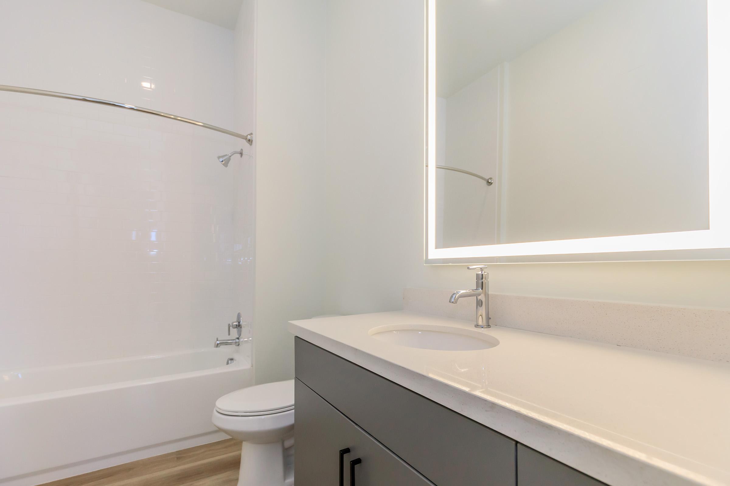 A modern bathroom featuring a clean design. The image shows a bathtub with a shower curtain, a sleek vanity with a circular mirror, and a contemporary faucet. The walls are painted light colors, and the flooring appears to be wooden. The space is well-lit and minimalistic.