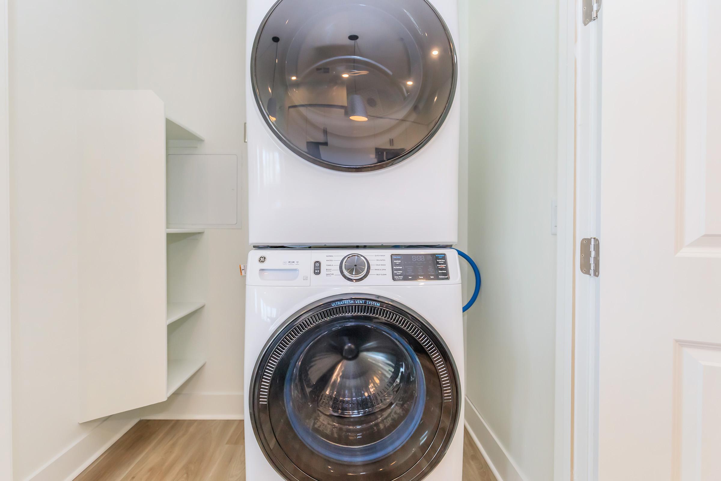A modern, stacked washer and dryer in a clean laundry room. The washer is on the bottom with a glass door revealing the drum, while the dryer is on top, also with a glass door. The surrounding area features light-colored walls and a shelf unit to the left.