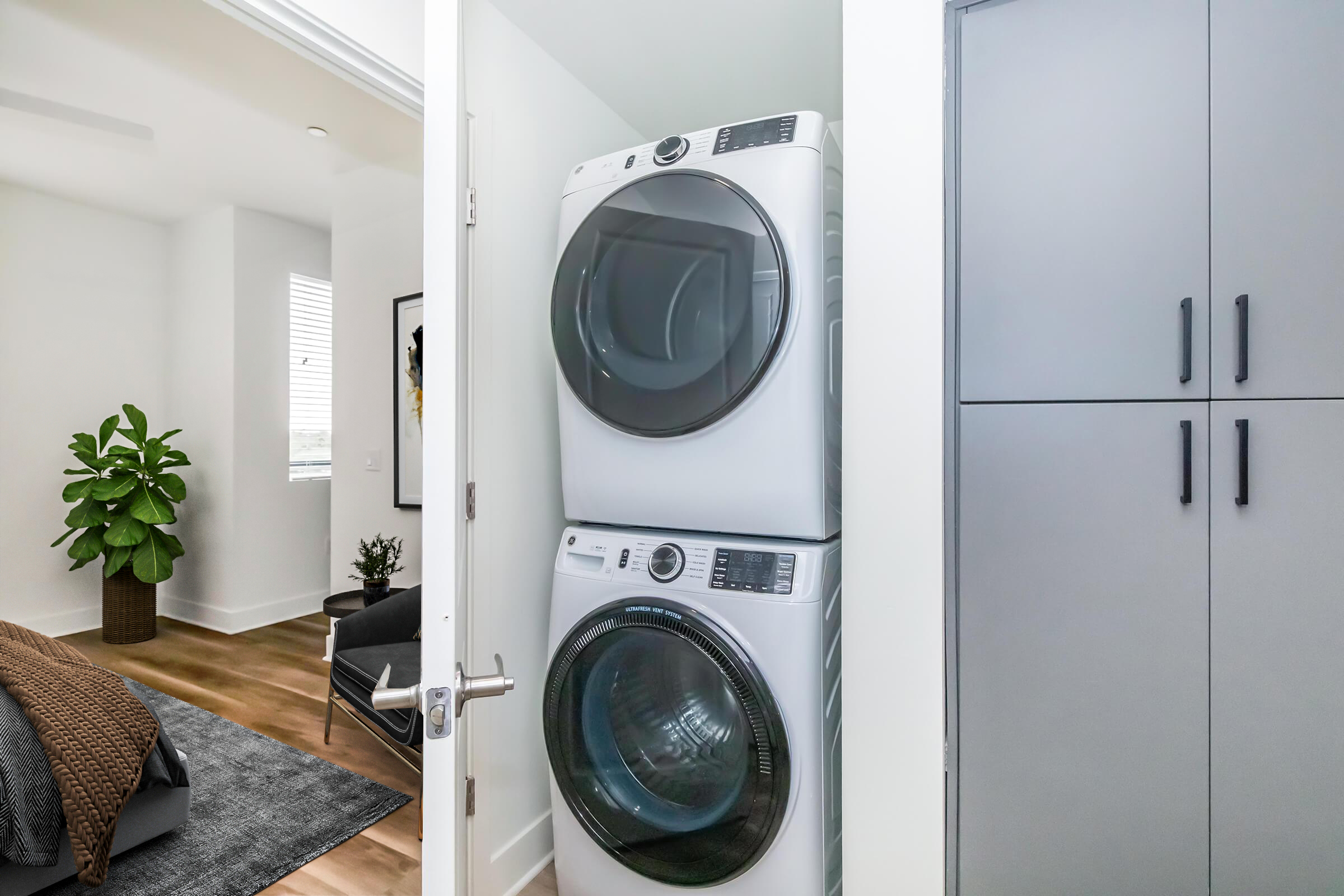 A modern laundry area featuring a stacked washer and dryer in a well-lit corner. The background includes a potted plant and hints of a cozy bedroom with a neatly made bed, light wood flooring, and contemporary cabinetry.