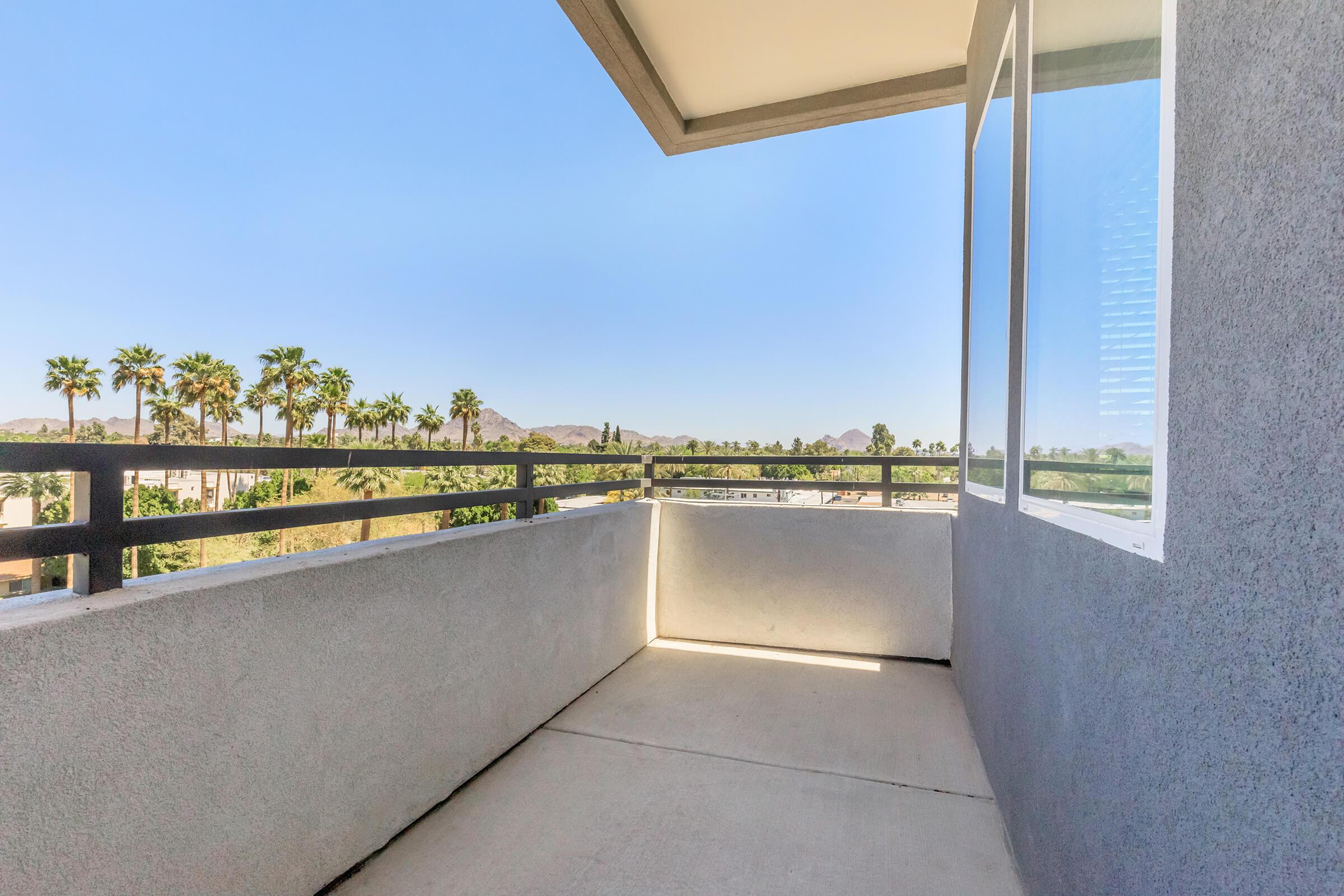 A spacious, empty balcony with a concrete floor and gray walls. The view features palm trees and distant mountains under a clear blue sky, highlighting a serene outdoor space for relaxation.