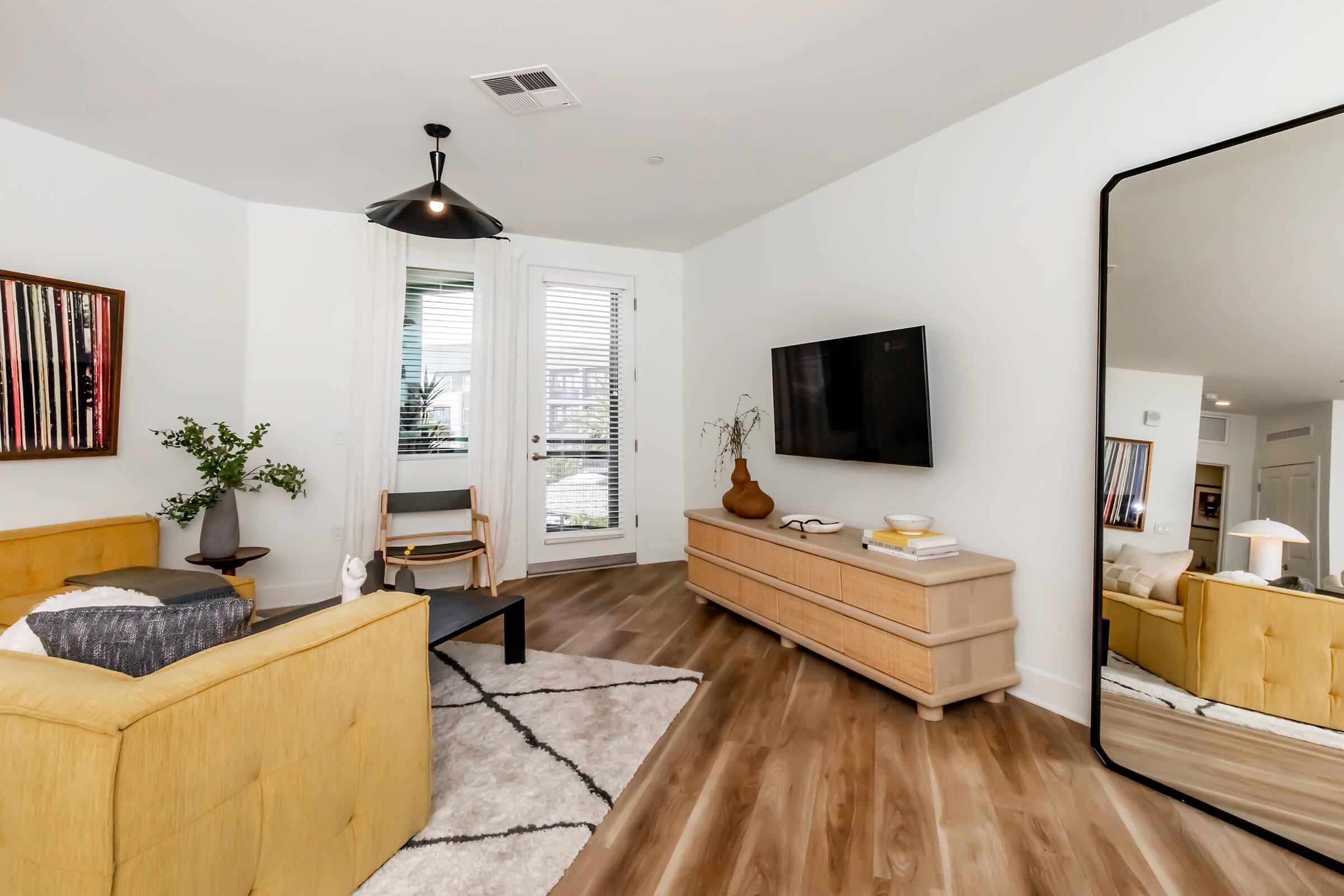 Modern living room featuring a yellow sofa, a black accent chair, and a wooden media console. A large mirror reflects the space, with a flat-screen TV mounted on the wall. Natural light filters through white curtains, and decorative plants and art pieces add a stylish touch to the cozy atmosphere.