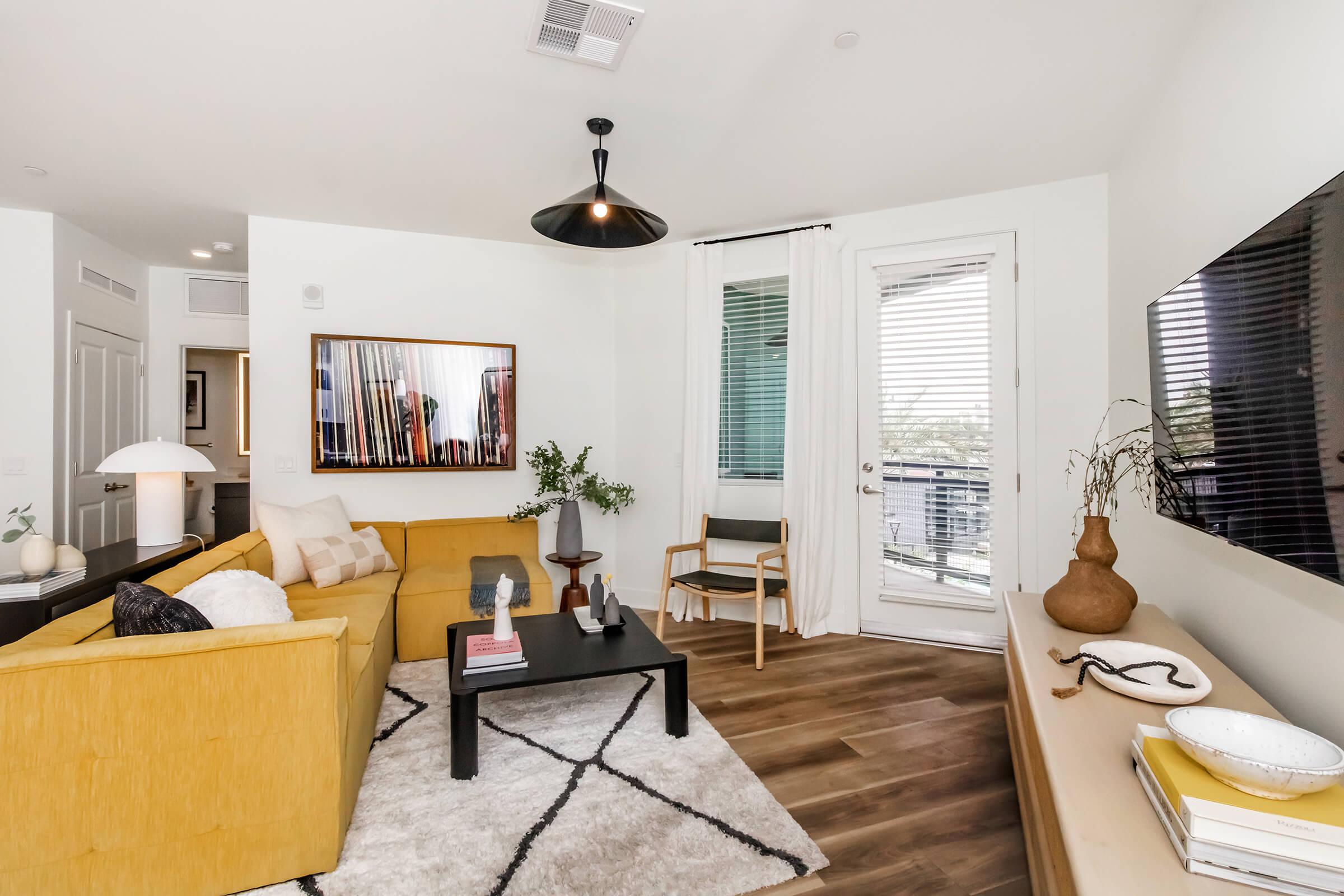 A modern living room featuring a yellow sofa, a black coffee table, and a flat-screen TV mounted on the wall. A large window with curtains allows natural light, and a wooden chair is positioned nearby. Decorative items include a potted plant and a vase on the table, creating a cozy and inviting atmosphere.