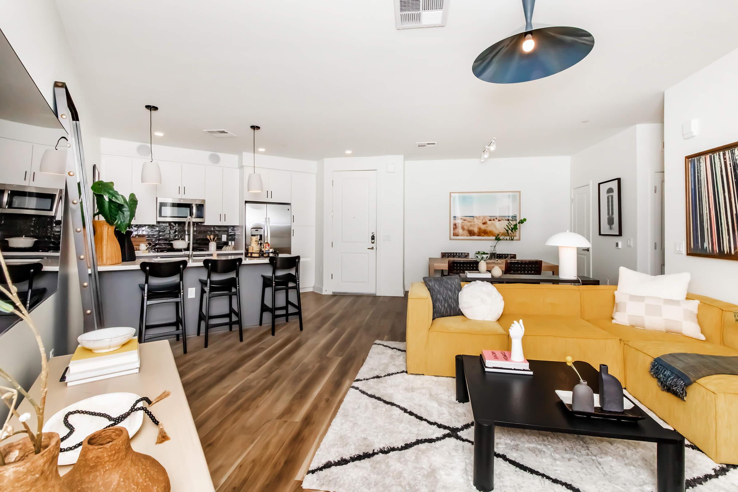 A modern living space featuring a bright yellow couch, a black coffee table, and a stylish kitchen in the background with stainless steel appliances. The room has a cozy vibe with decorative elements like a vase, artwork on the walls, and a light fixture overhead.