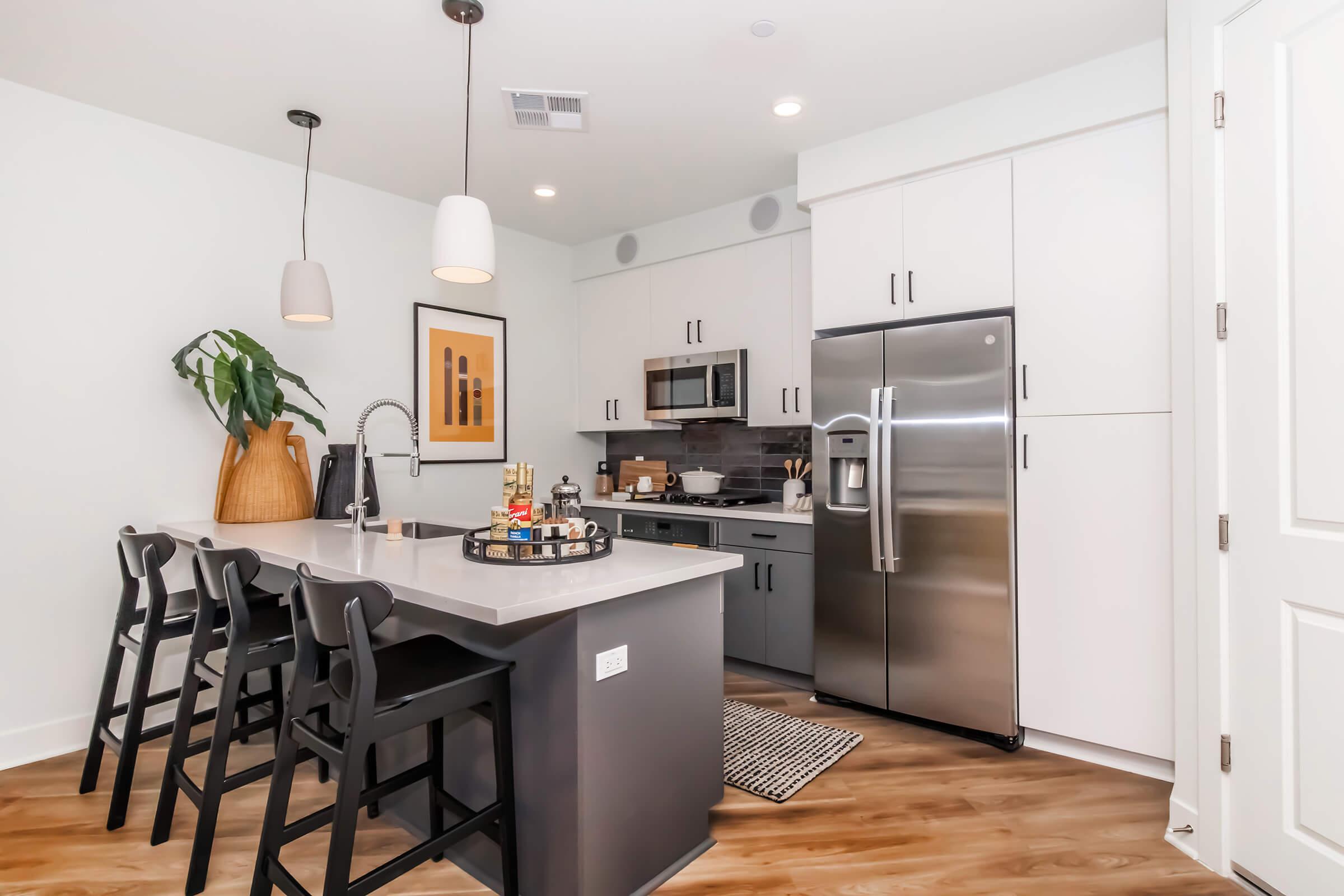 Modern kitchen with a sleek design featuring stainless steel appliances, including a refrigerator and microwave. A central island with seating for four is highlighted by stylish pendant lighting. A decorative plant and framed artwork add a touch of warmth to the minimalist space, complemented by a patterned rug on the wood floor.