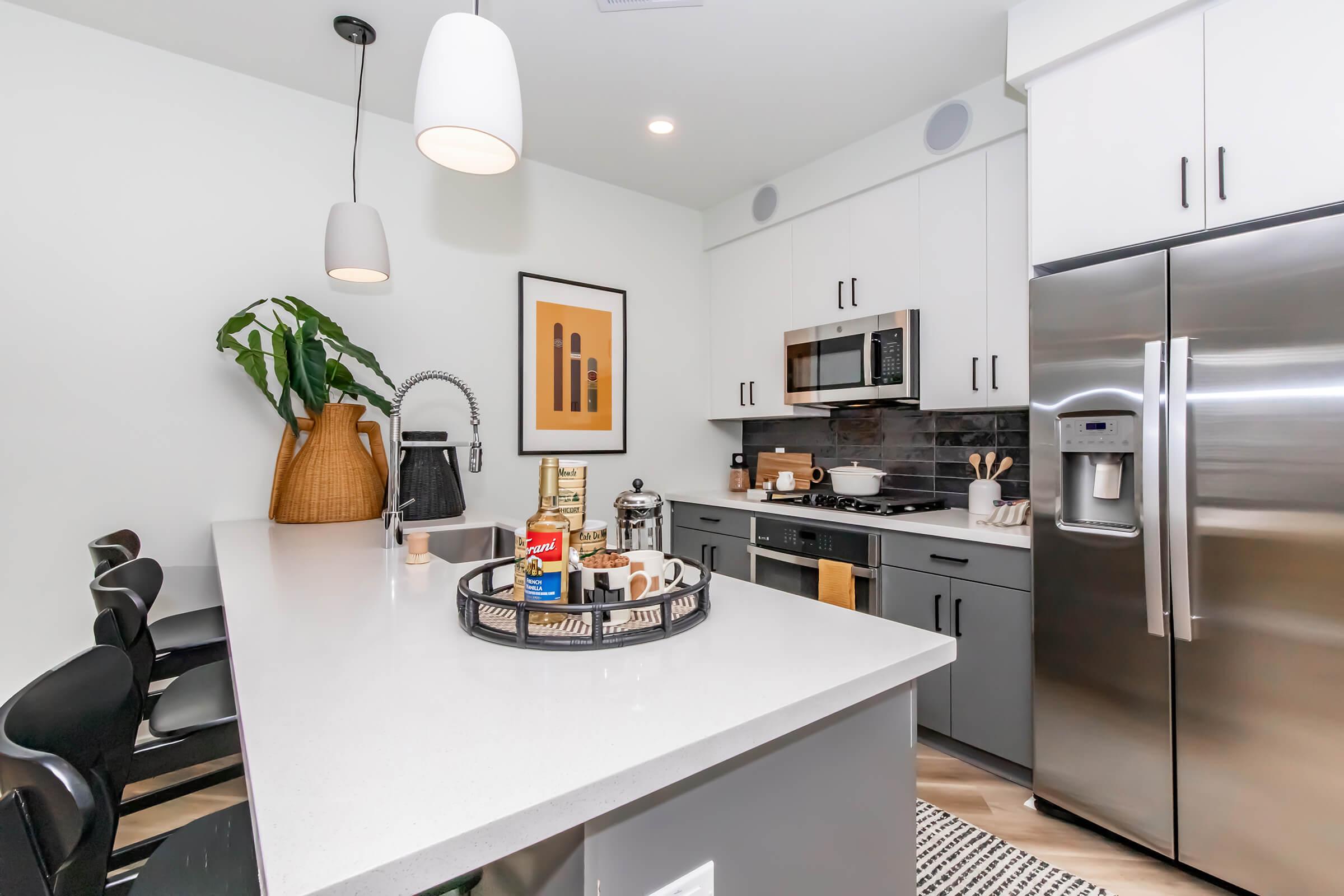 Modern kitchen featuring a light gray and white color scheme. The space includes stainless steel appliances, a large island with bar stools, and decorative items on the counter. A wall-mounted art piece and a potted plant add a touch of style to the contemporary design. Natural light illuminates the area.