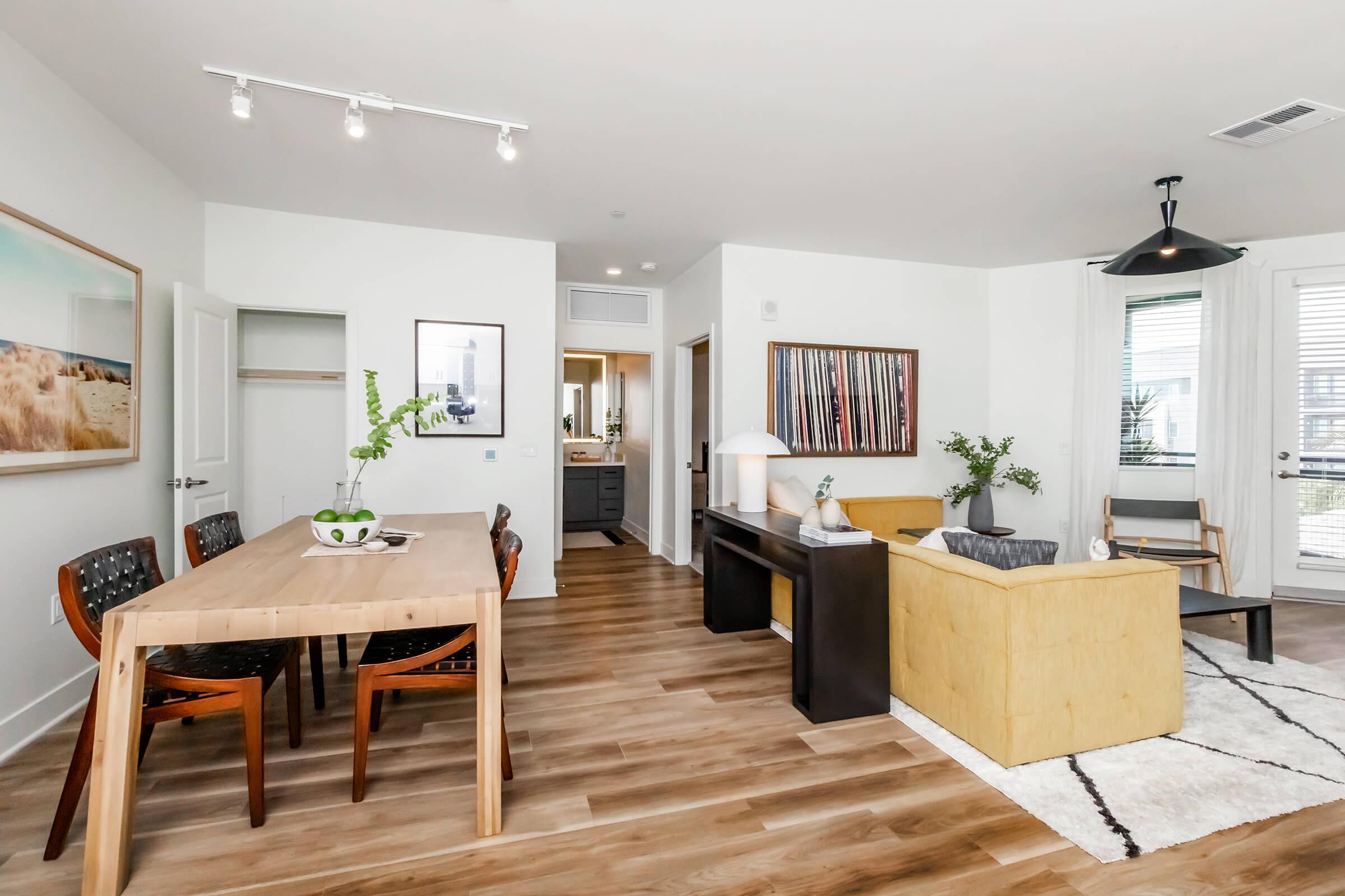A modern interior space featuring a dining area with a wooden table and chairs, a cozy living room with a yellow sofa and a black lamp, and artwork on the walls. Natural light fills the room through large windows, creating a bright and inviting atmosphere.